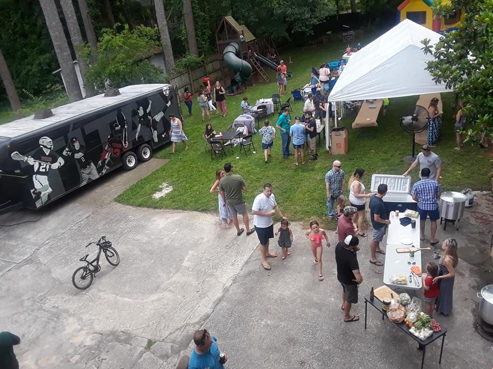 Outdoor backyard gathering with numerous people, a food truck, children, a large tent, and a playset in a wooded area.