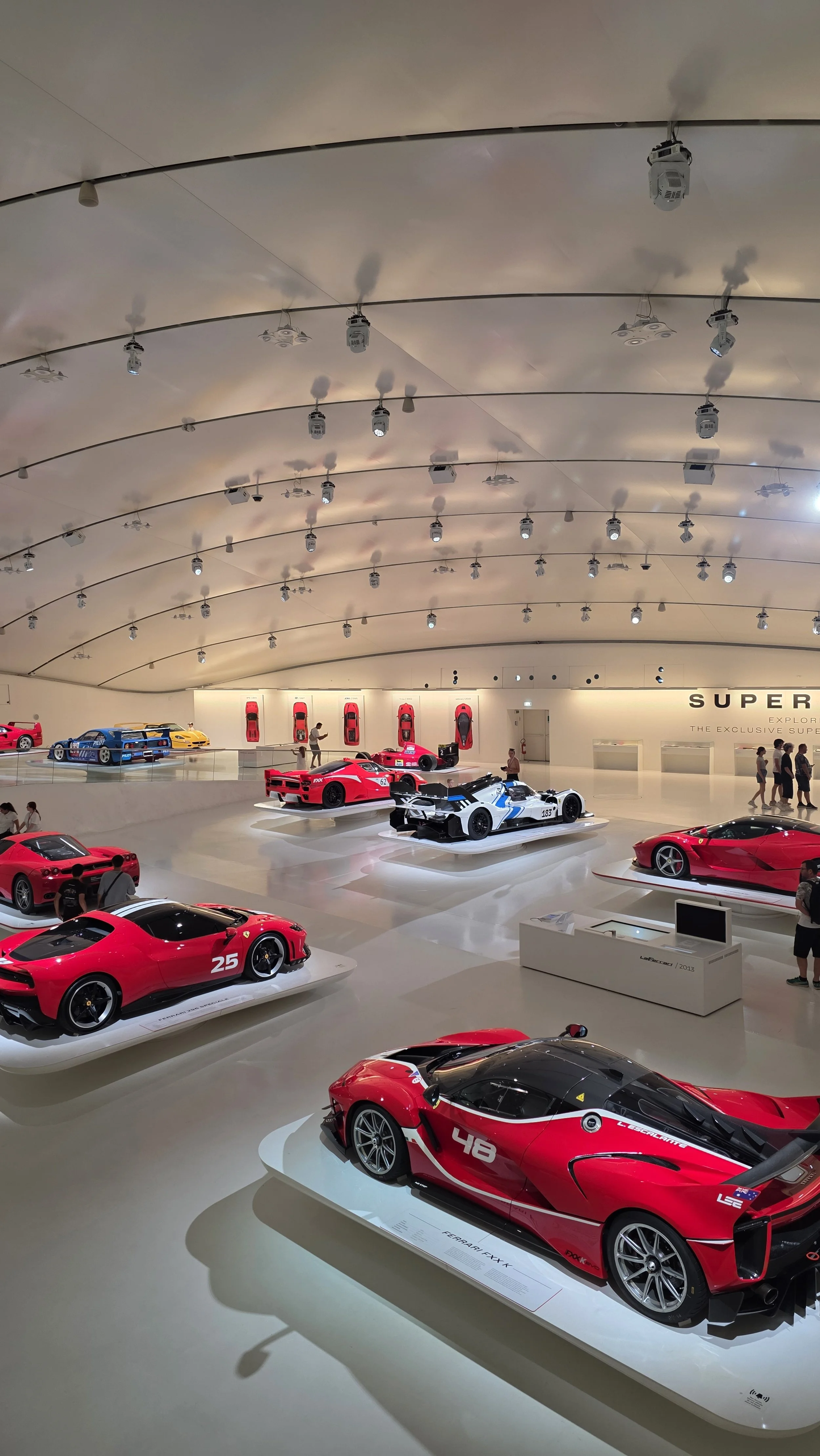 A car museum with a collection of red and white race cars displayed on white platforms. The ceiling is curved with multiple spotlights, and visitors are walking around, observing the cars.