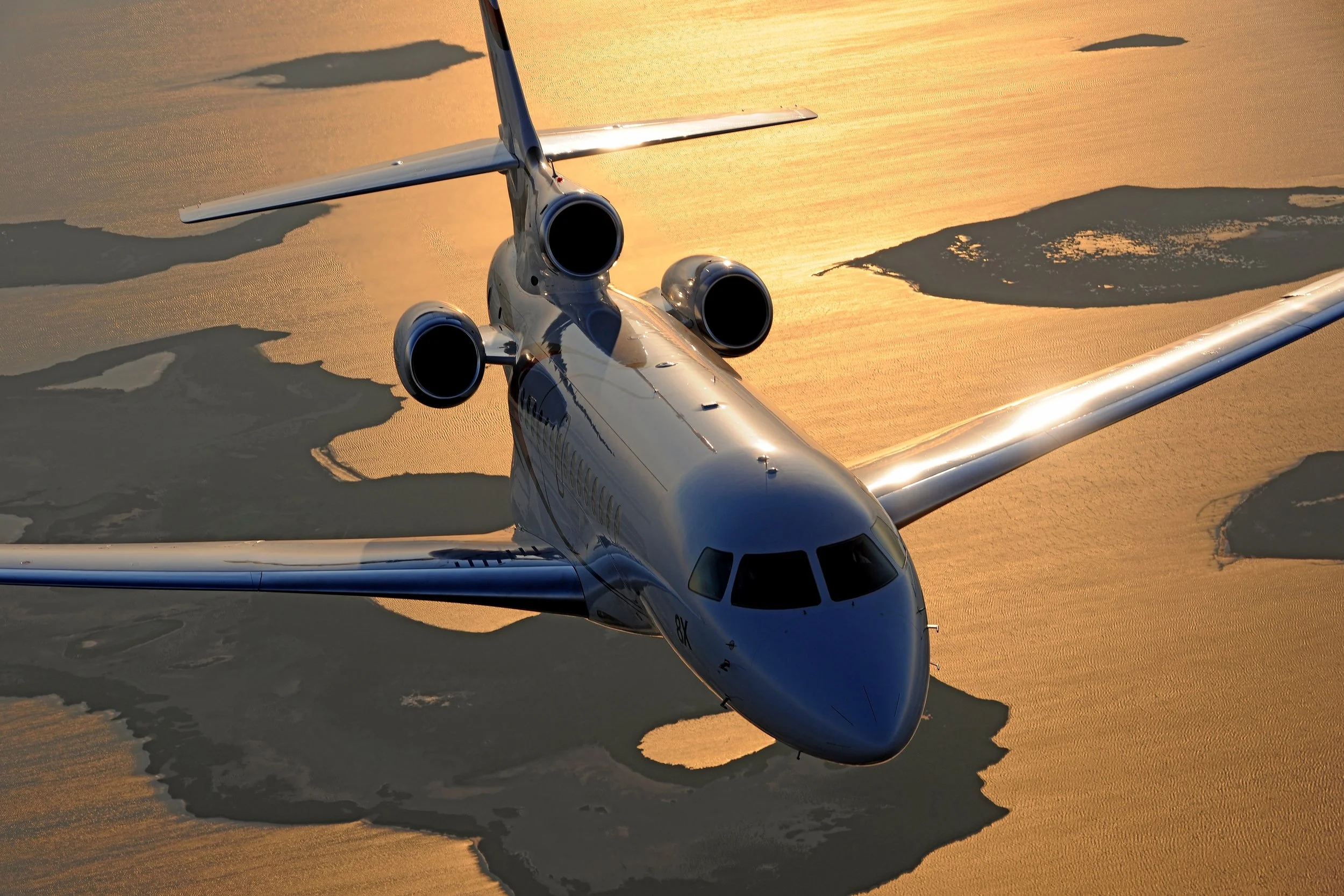 A silver private jet flying over a body of water during sunset with golden hues and small islands underneath.