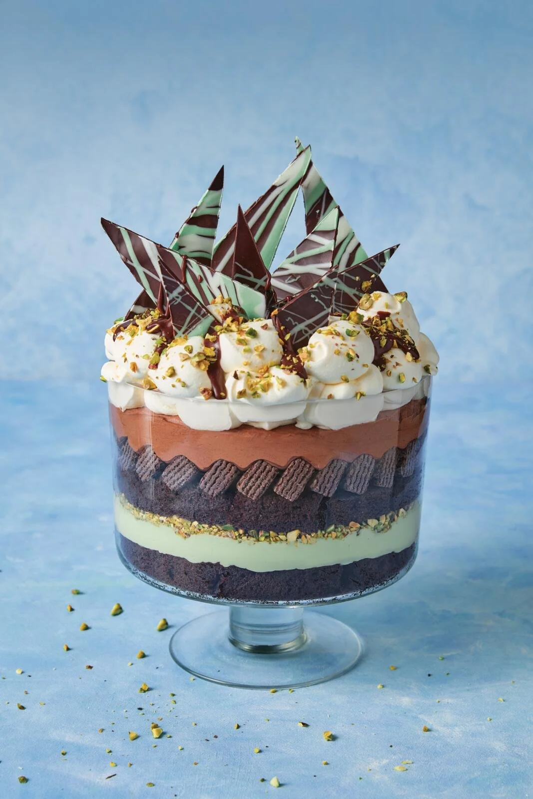 Layered chocolate dessert in a glass, topped with whipped cream, sliced chocolate, and chopped pistachios on a blue background.