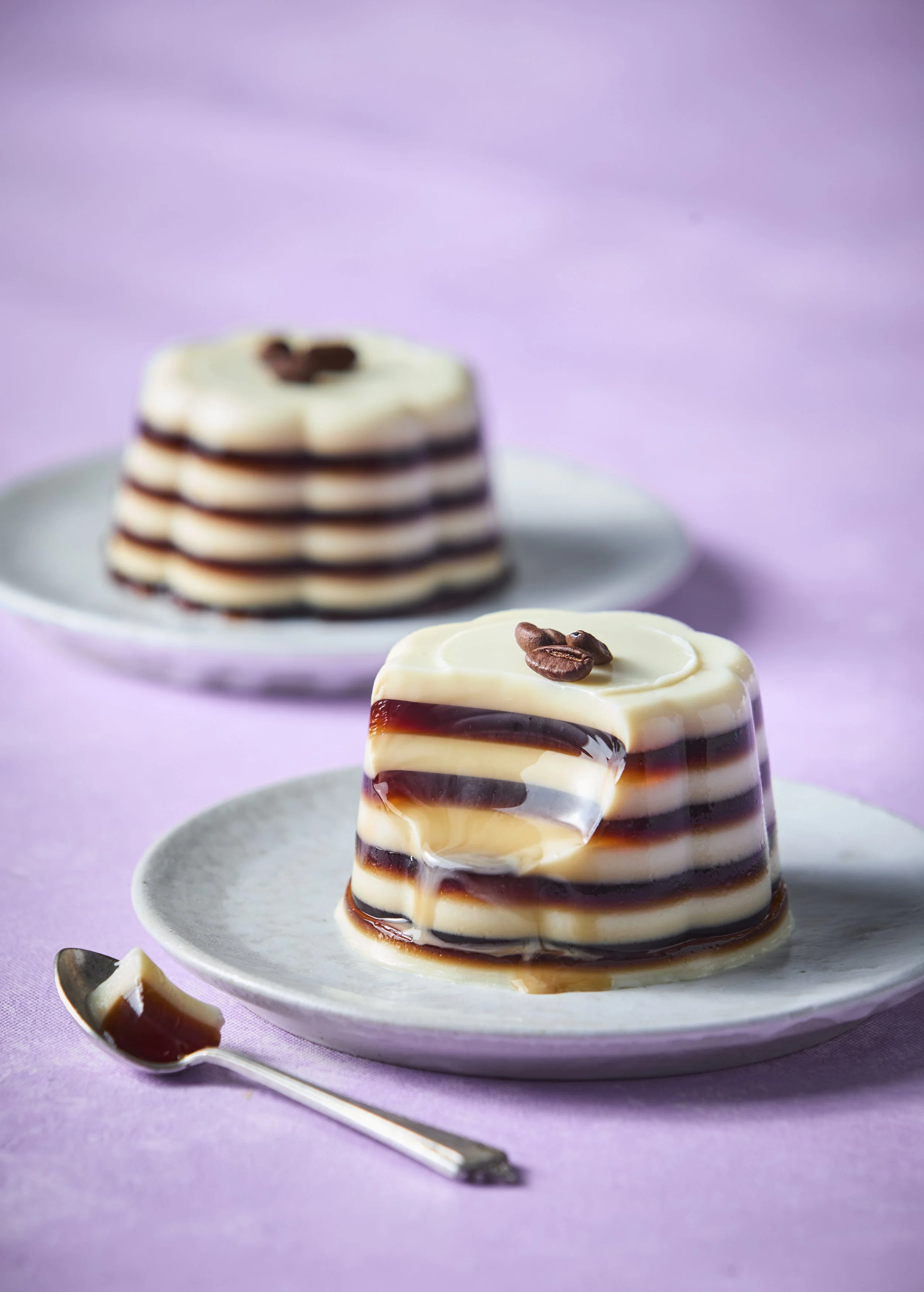 Two plates with layered panna cotta desserts topped with coffee beans on a lilac surface.