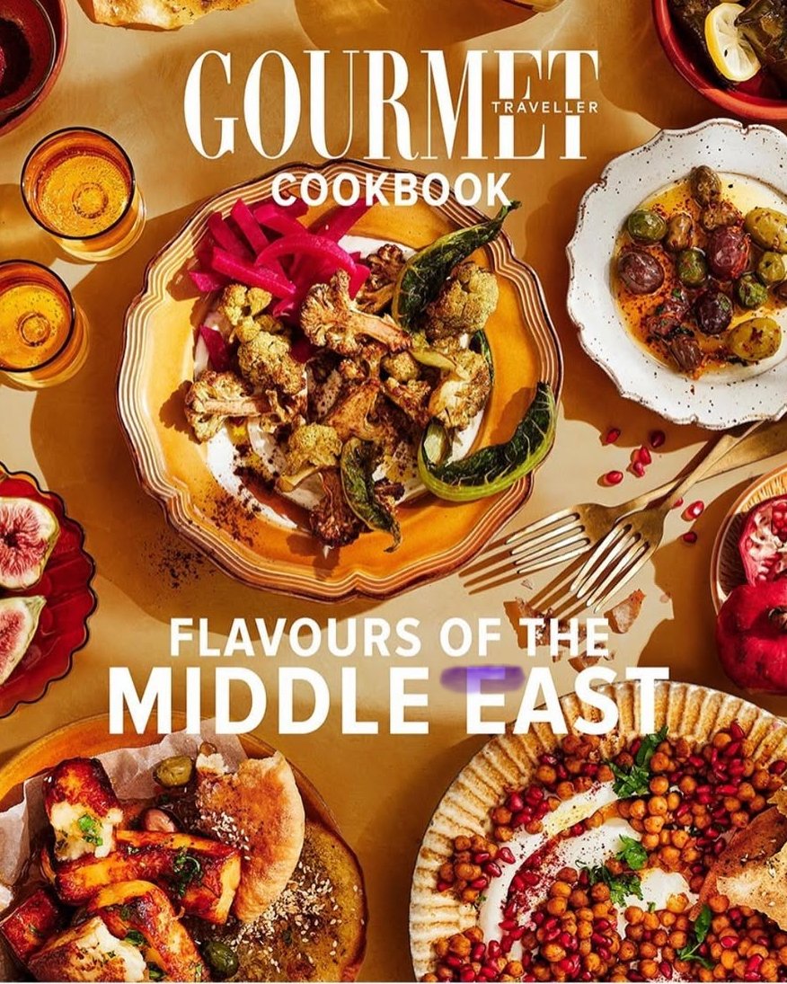 Table with Middle Eastern dishes including roasted vegetables, stuffed grape leaves, pomegranate seeds, and olives, with small glasses of tea or juice.