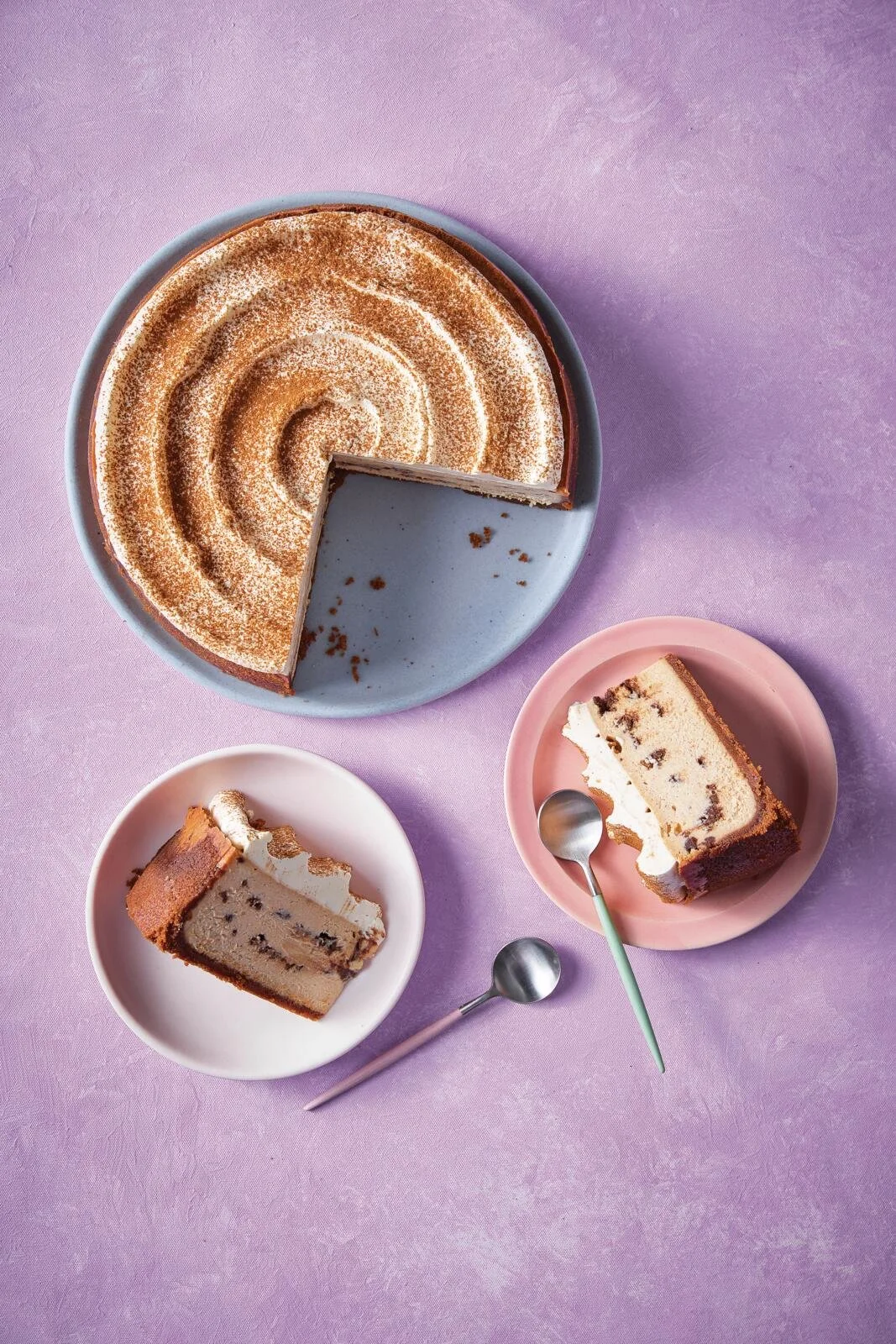 Top view of a large round cake with a slice cut out, placed on a blue plate. Two slices of cake on small pink and white plates with spoons, set on a purple surface.