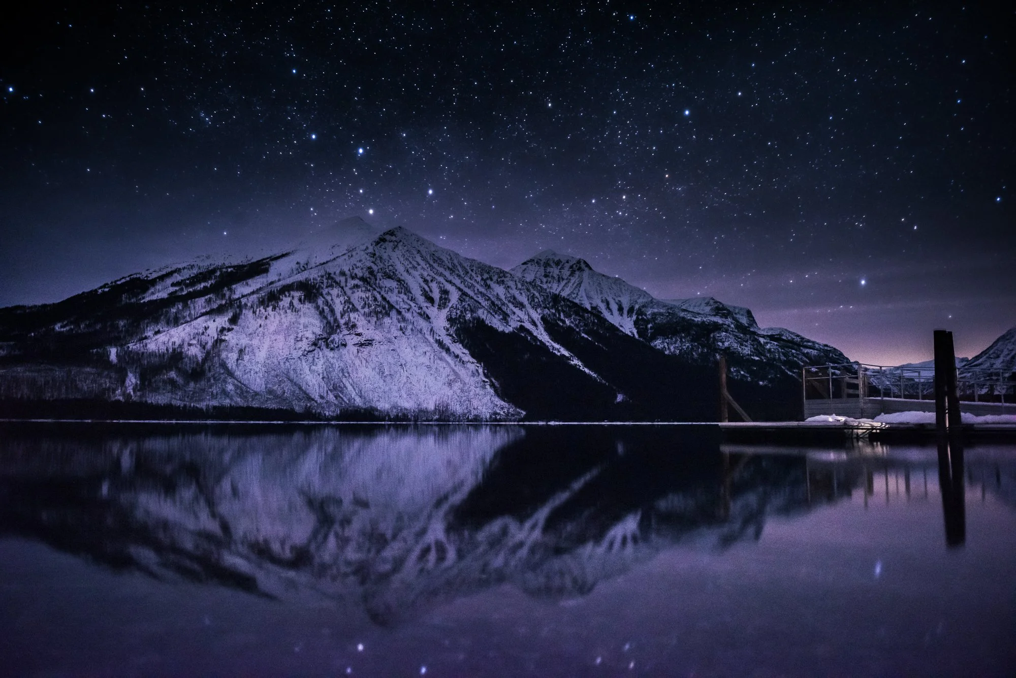 Nighttime view of snow-covered mountains reflected in a calm lake under a star-filled sky.