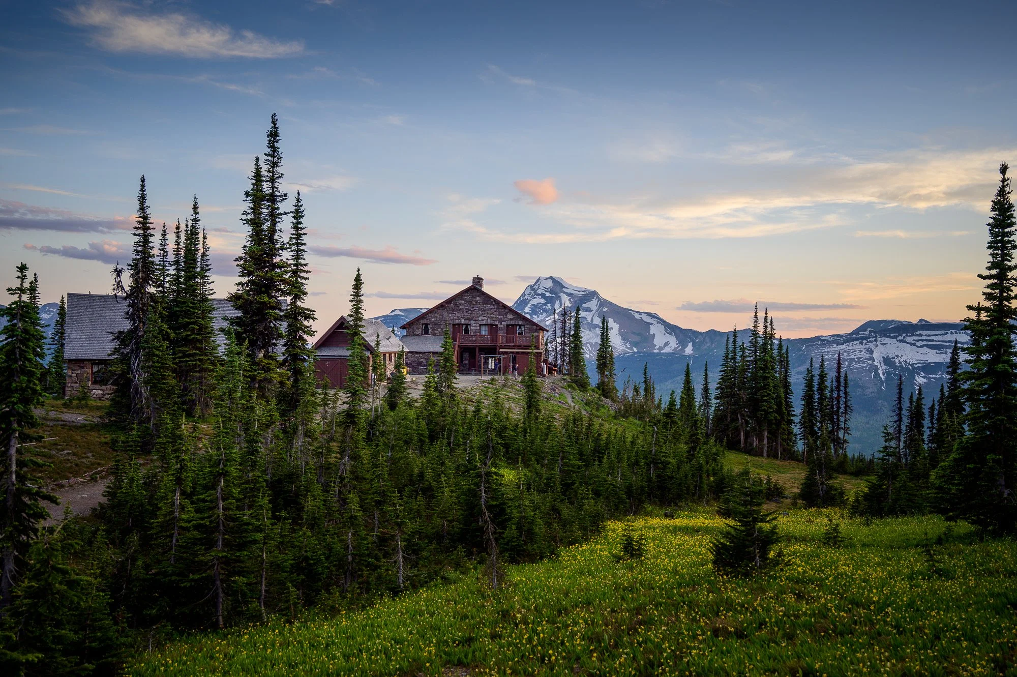 Mountain cabin surrounded by pine trees with a snow-capped mountain peak in the background, during sunset.