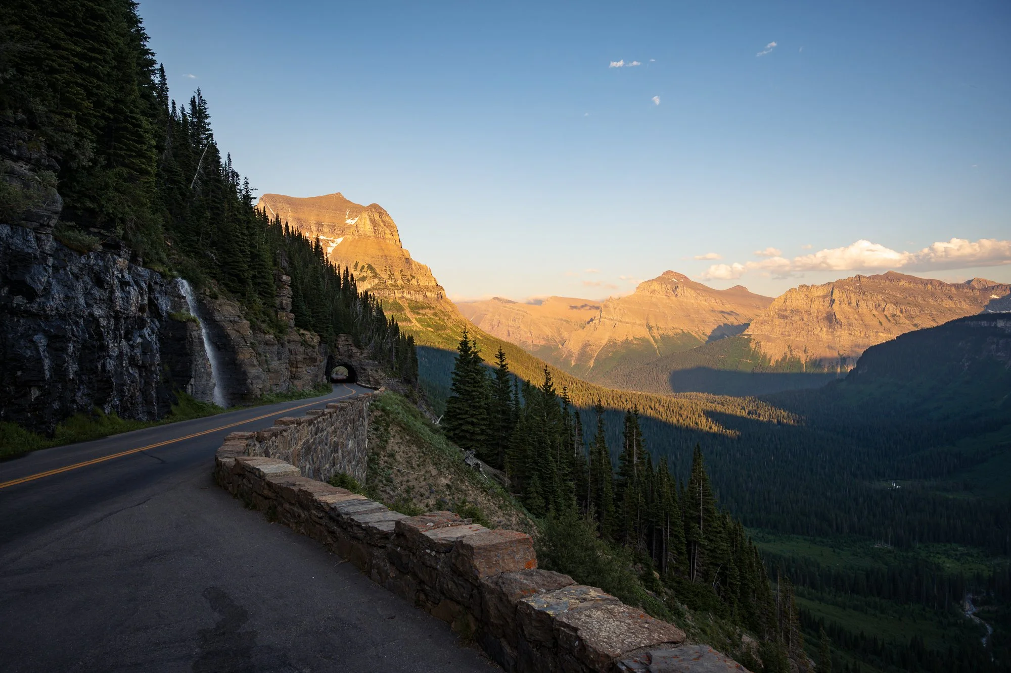 A winding road through a mountainous landscape with evergreen trees, cliffs, and a waterfall at sunset.
