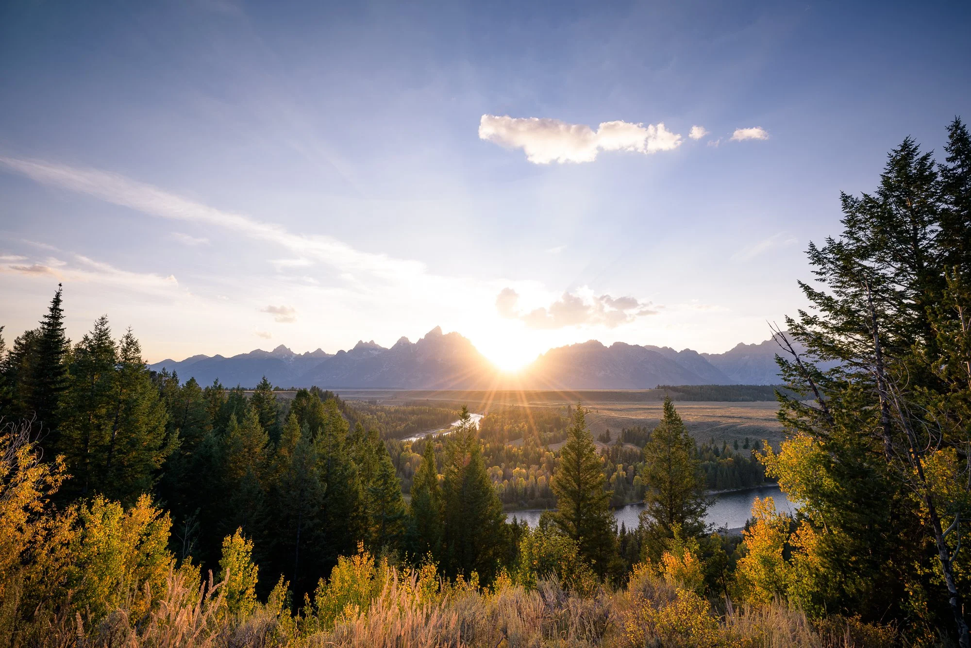 Sunrise over a mountain range with a river and a forest of green and yellow trees in the foreground.
