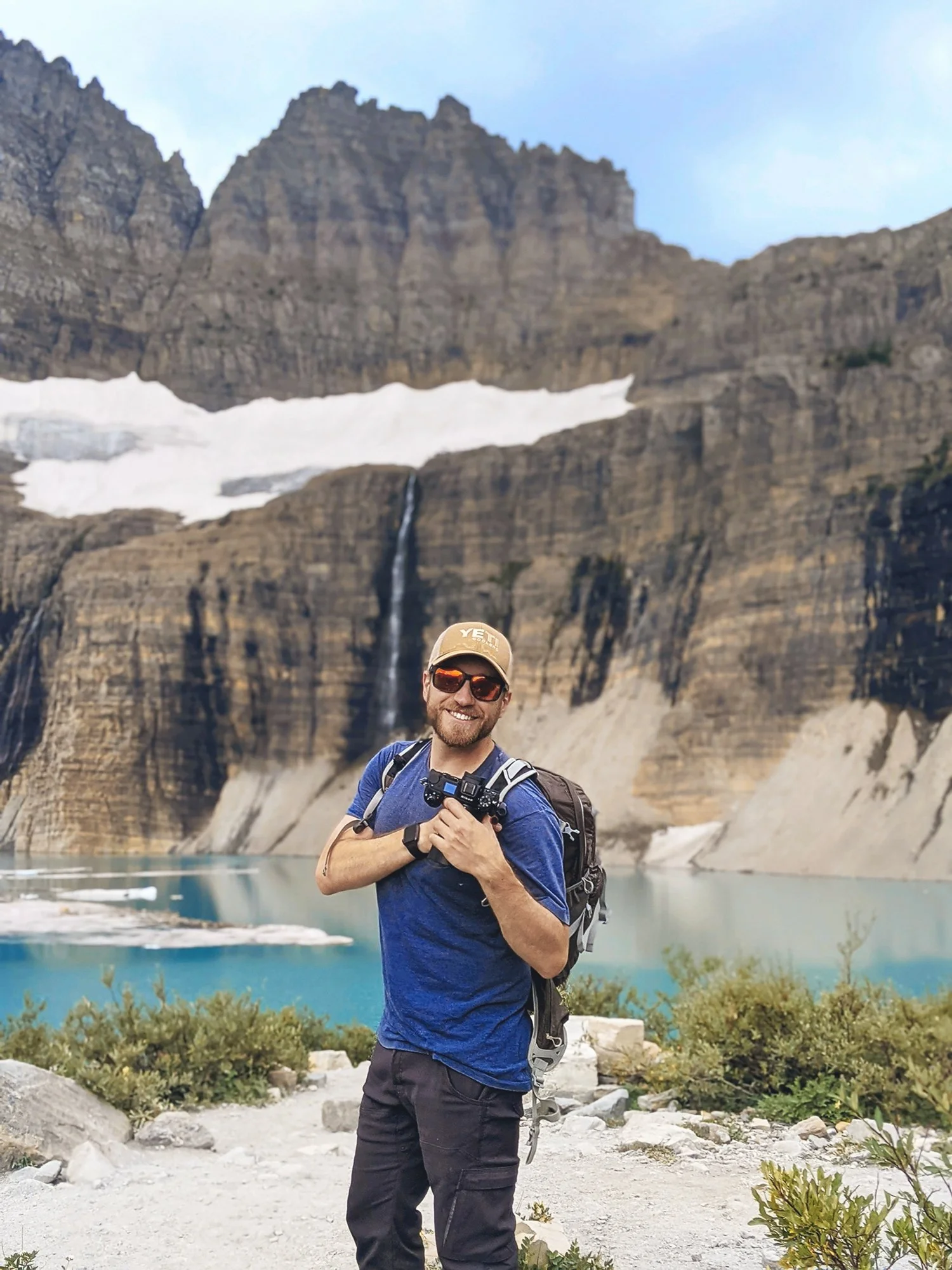 A man smiling and holding a camera in front of a mountain landscape with a lake, waterfall, and snow patches.