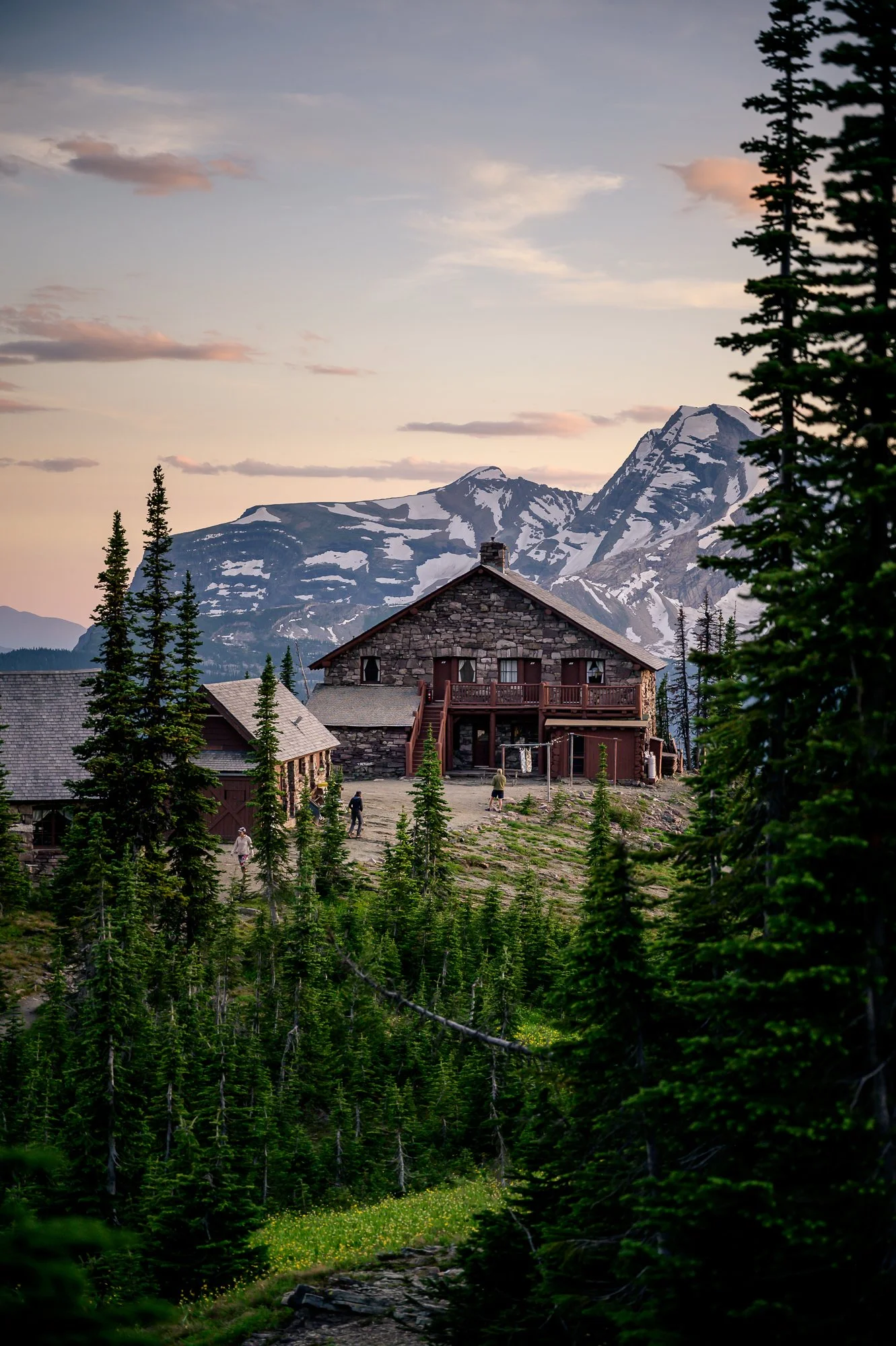 A scenic mountain landscape with snow-capped peaks, a stone cabin with a wooden deck, surrounded by green pine trees, during sunset or sunrise.
