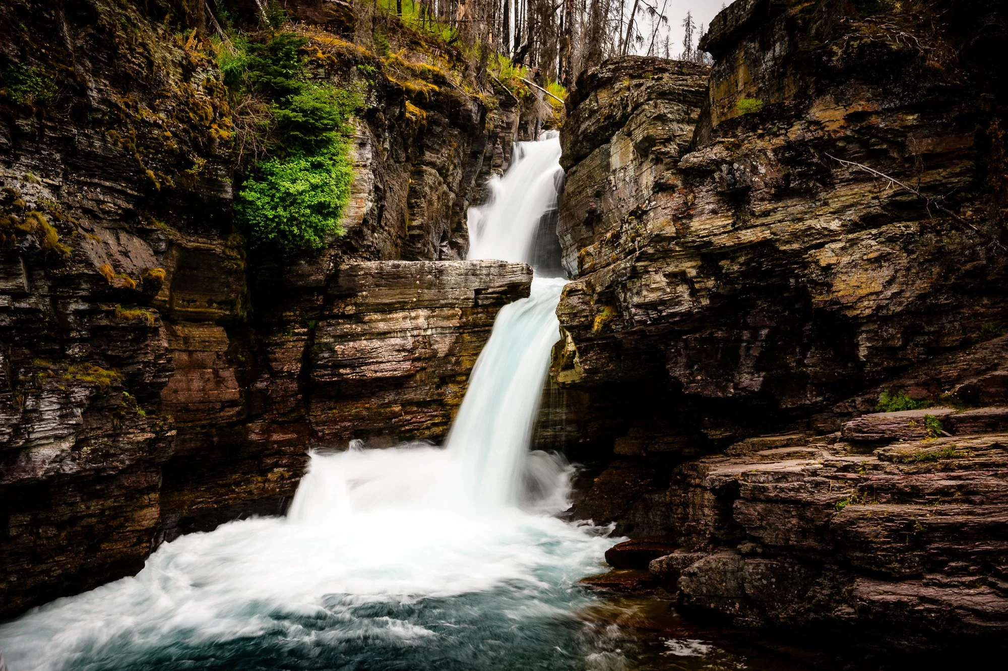 A waterfall cascading down rocky cliffs in a forested area, with green vegetation on the cliffs and water flowing into a river below.