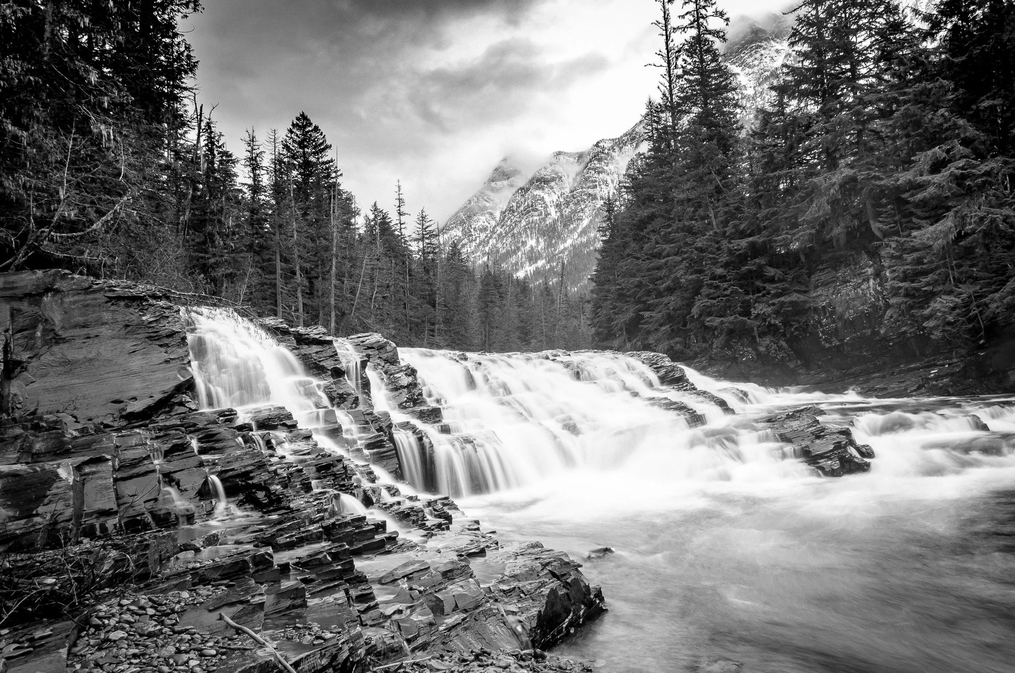 Black and white photo of a waterfall flowing over rocky terrain surrounded by tall evergreen trees, with snow-covered mountains in the background and cloudy sky above.