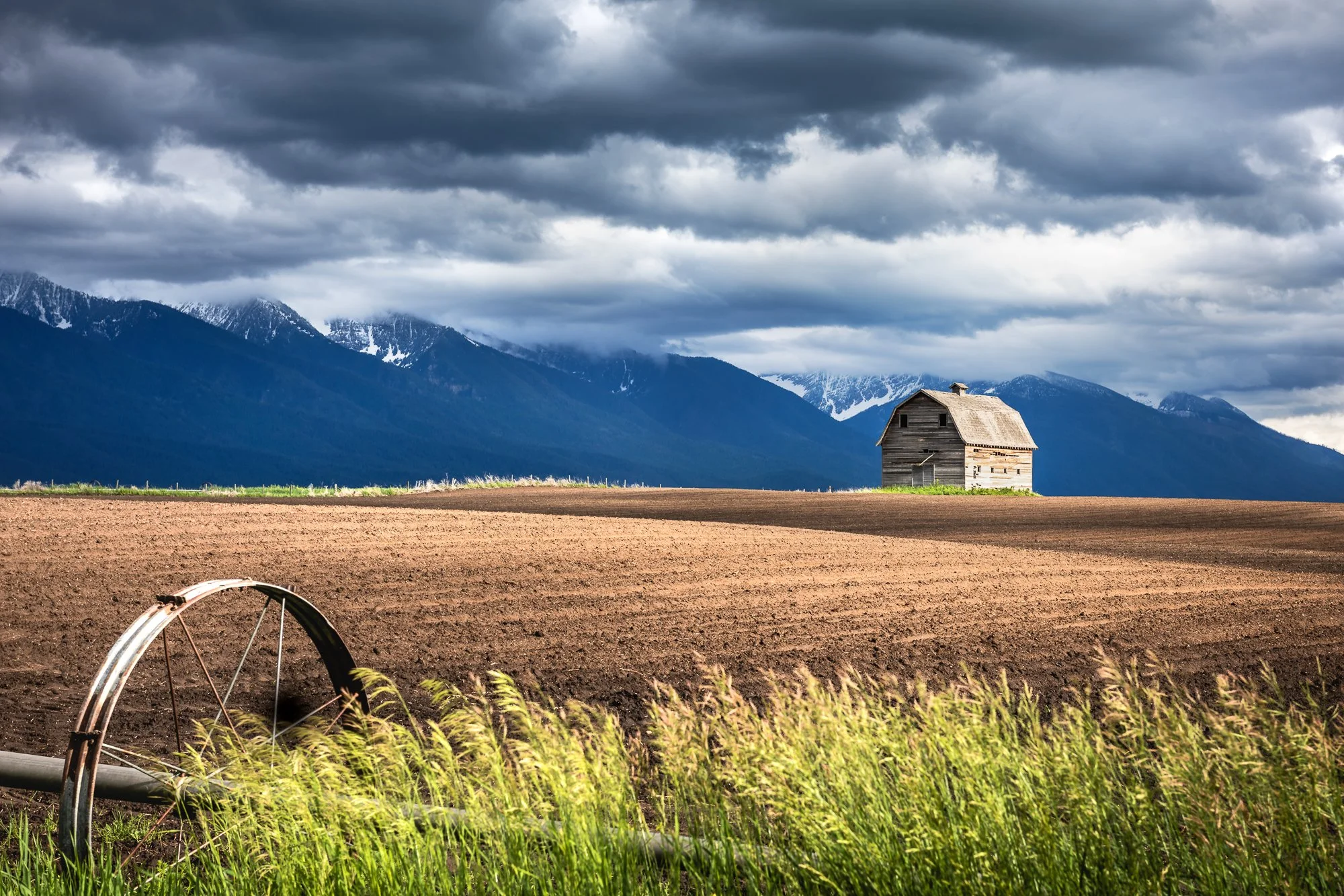 Scene of farmland with tilled soil, an old wooden barn, tall grass in the foreground, mountains in the background, and a sky filled with dark clouds.