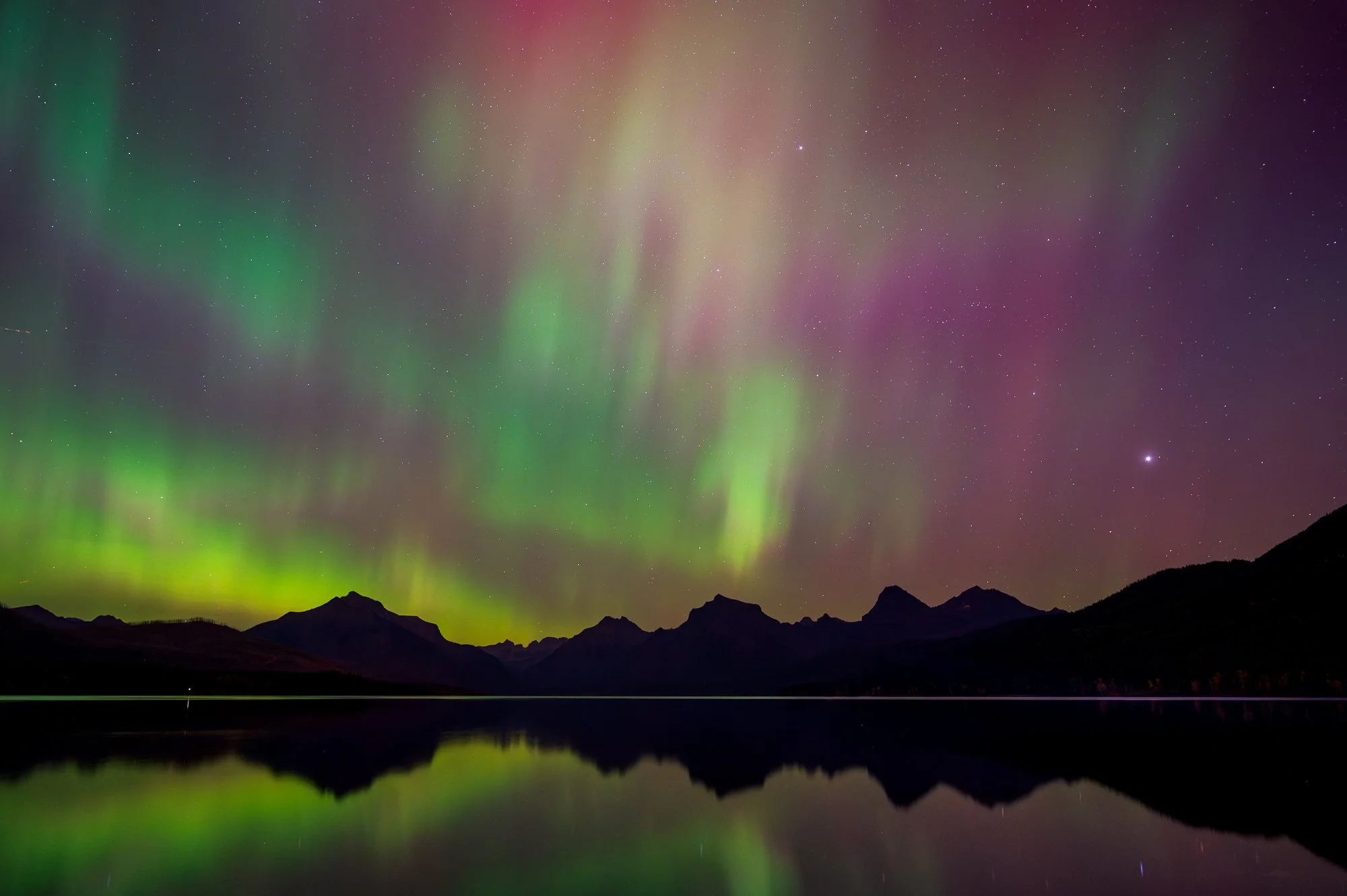 Northern lights (aurora borealis) over mountains and a calm lake at night with starry sky.