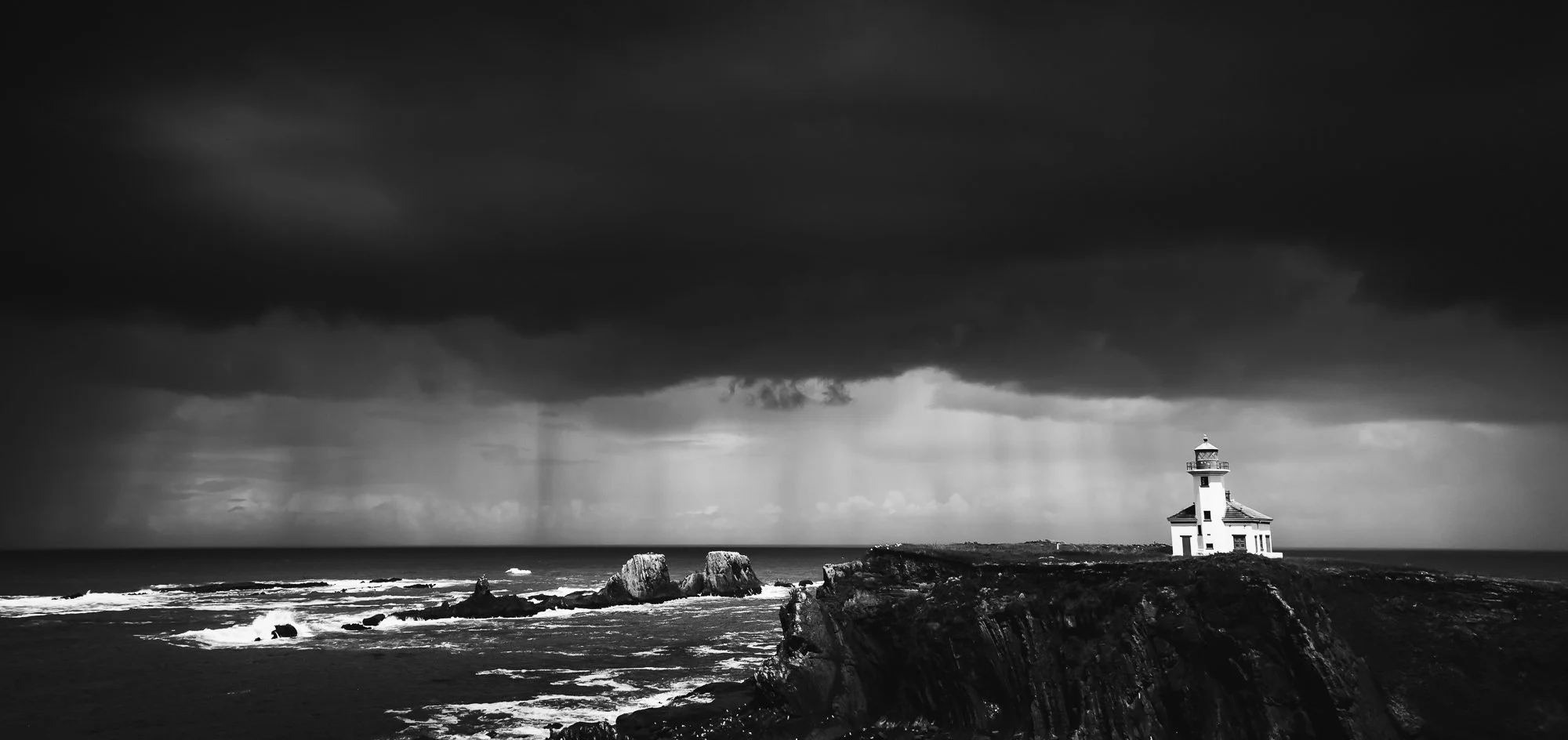 Black and white photograph of a lighthouse on a rocky cliff during a storm, with dark, heavy clouds overhead and rain falling over the ocean.