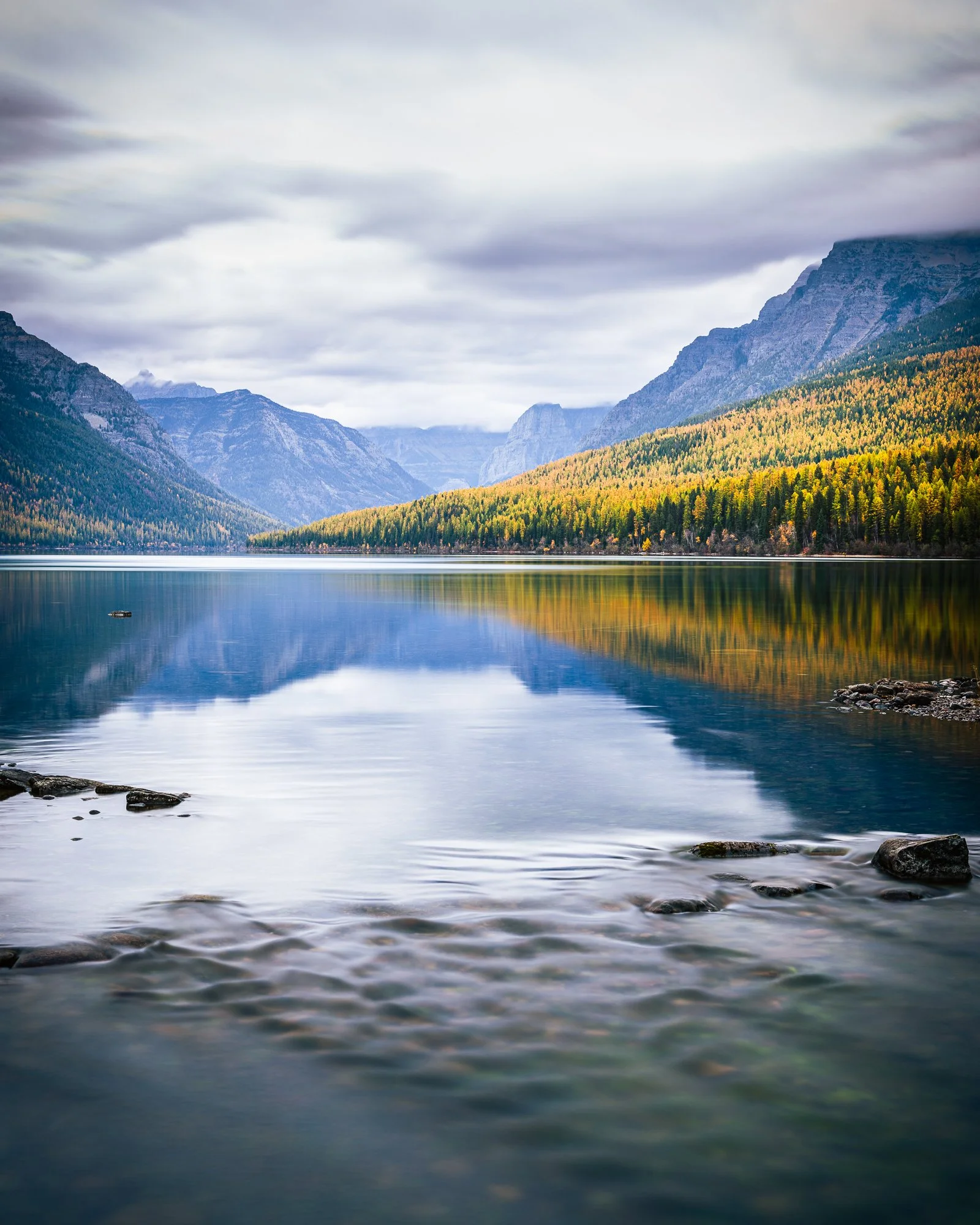 Scenic view of a calm lake surrounded by mountains and dense forest, with cloudy sky above.