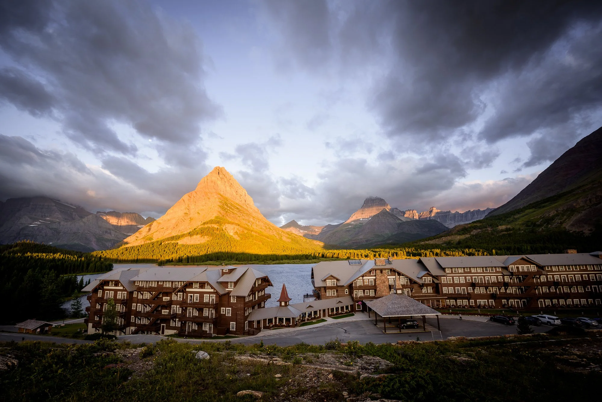 Large mountain with a pointed peak illuminated by sunlight, with other mountain peaks in the background, a river, and a large wooden hotel or lodge in the foreground under cloudy skies.