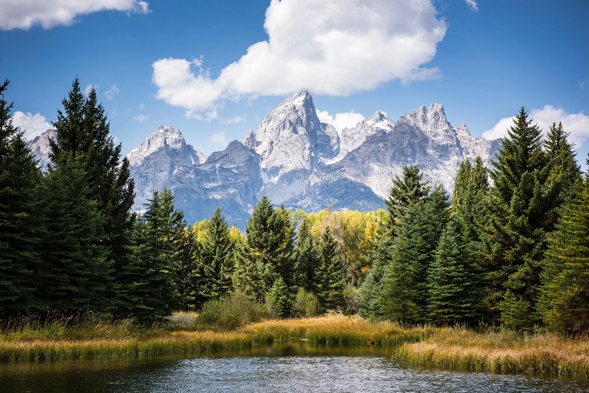 Scenic landscape of a pond surrounded by lush green trees, with tall, snowy mountain peaks in the background under a partly cloudy sky.