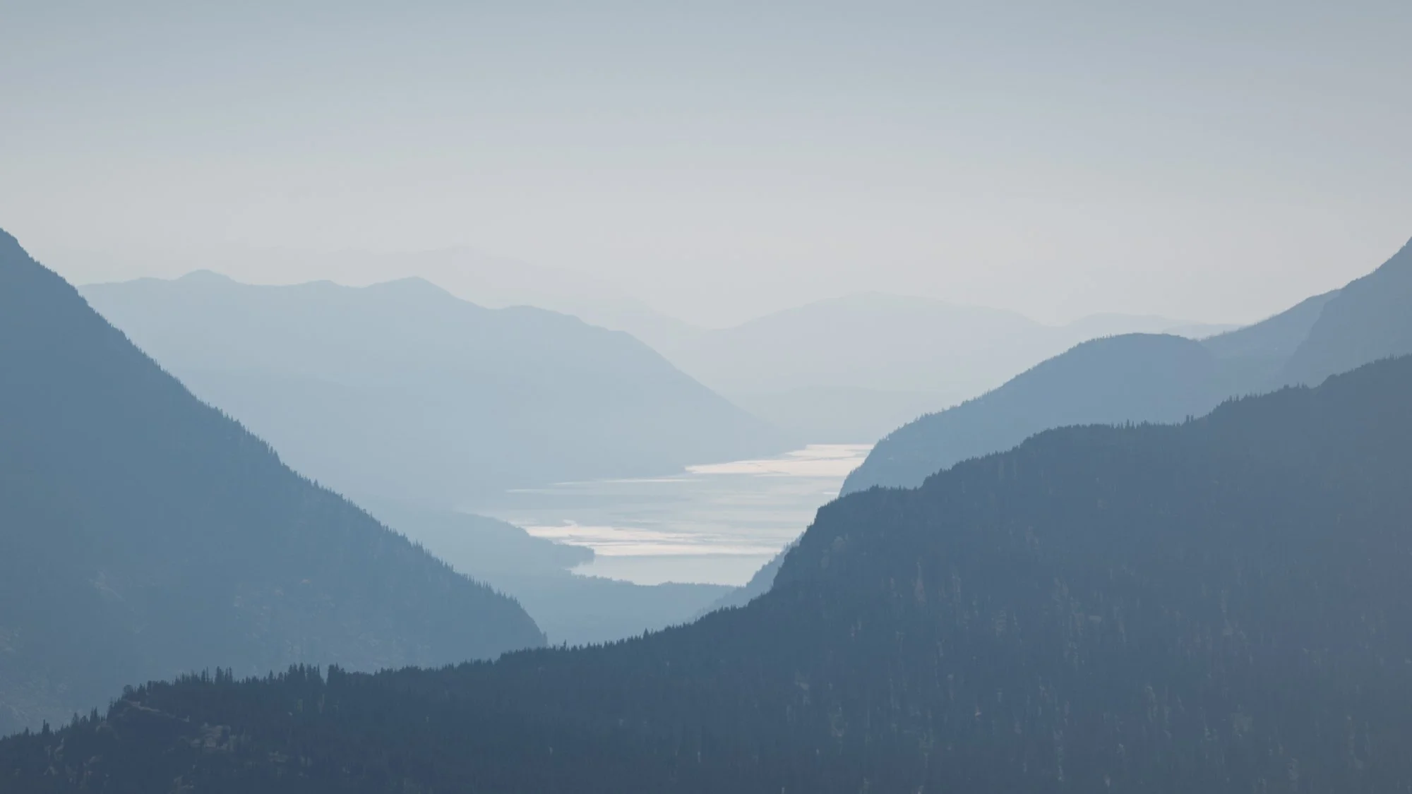 Mountain landscape with multiple ranges in layers, a river or lake in the distant valley, and a hazy sky.