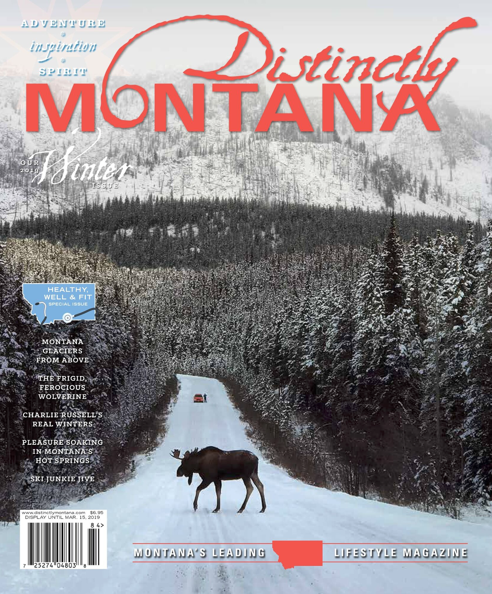 Cover of Montana lifestyle magazine featuring a snowy scene with a moose walking on a snow-covered road through a forest, with snow-covered mountains in the background and a vehicle and person visible in the distance.