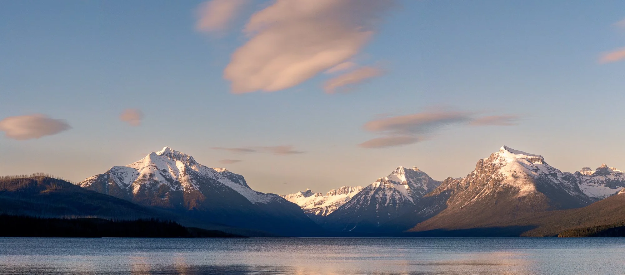 Snow-capped mountains reflecting in a calm lake under a partly cloudy sky during sunset.