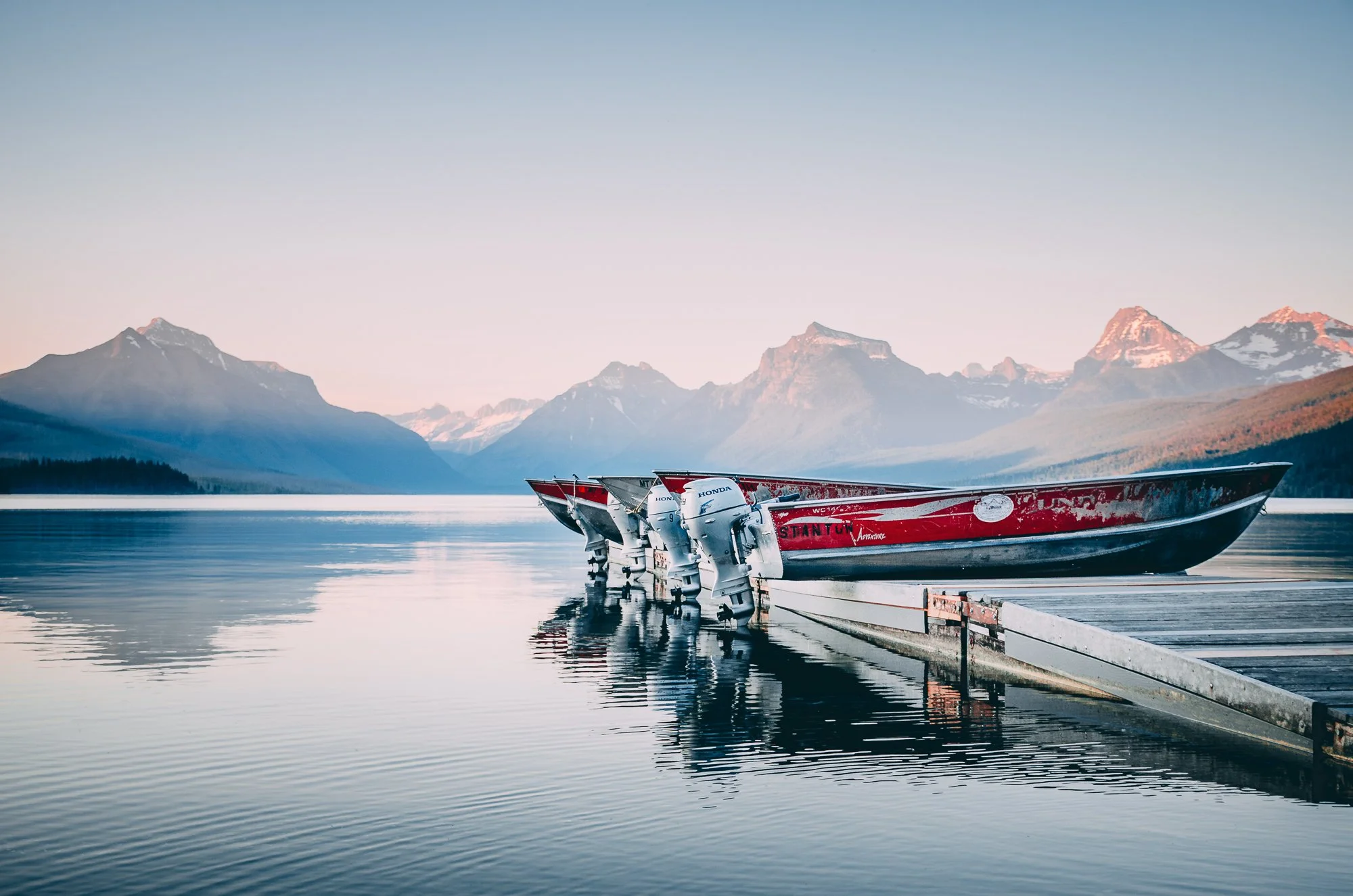Three speedboats with Honda engines docked at a wooden pier on a calm lake, with snow-capped mountains in the background during sunrise or sunset.