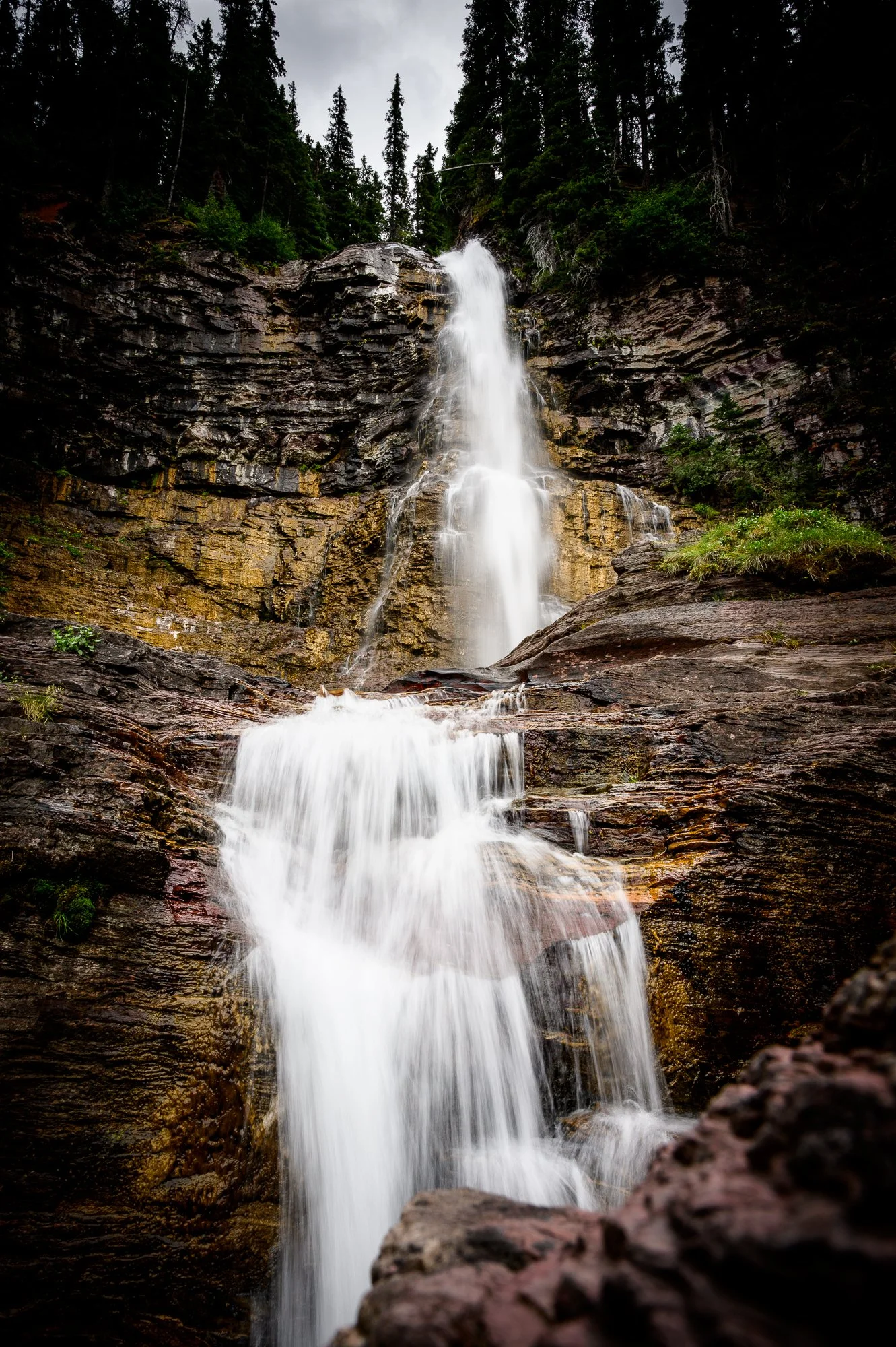 A tall waterfall cascading down a rocky cliff surrounded by dense green trees under an overcast sky.