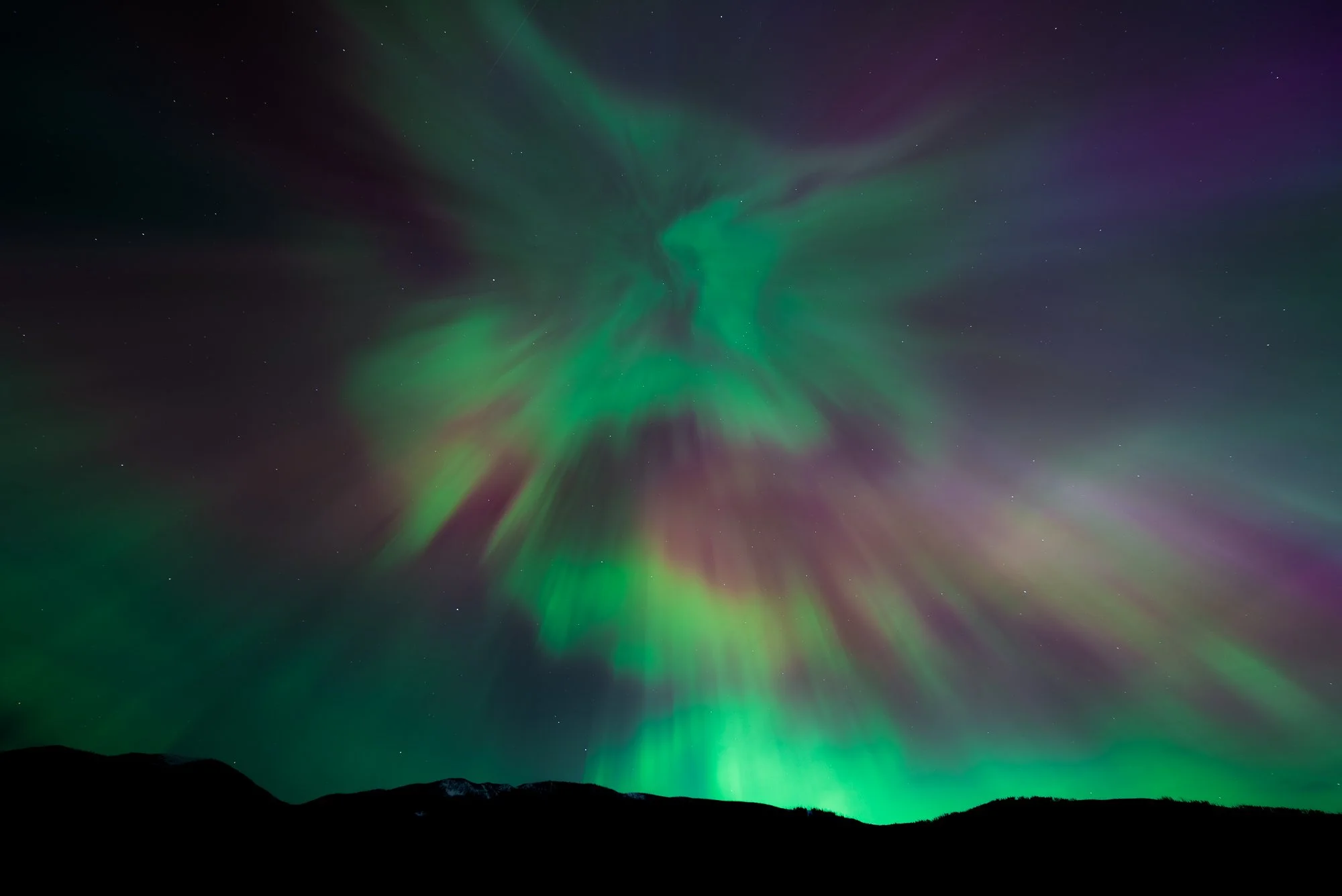 Northern lights display with swirling green and purple auroras over dark mountain silhouette