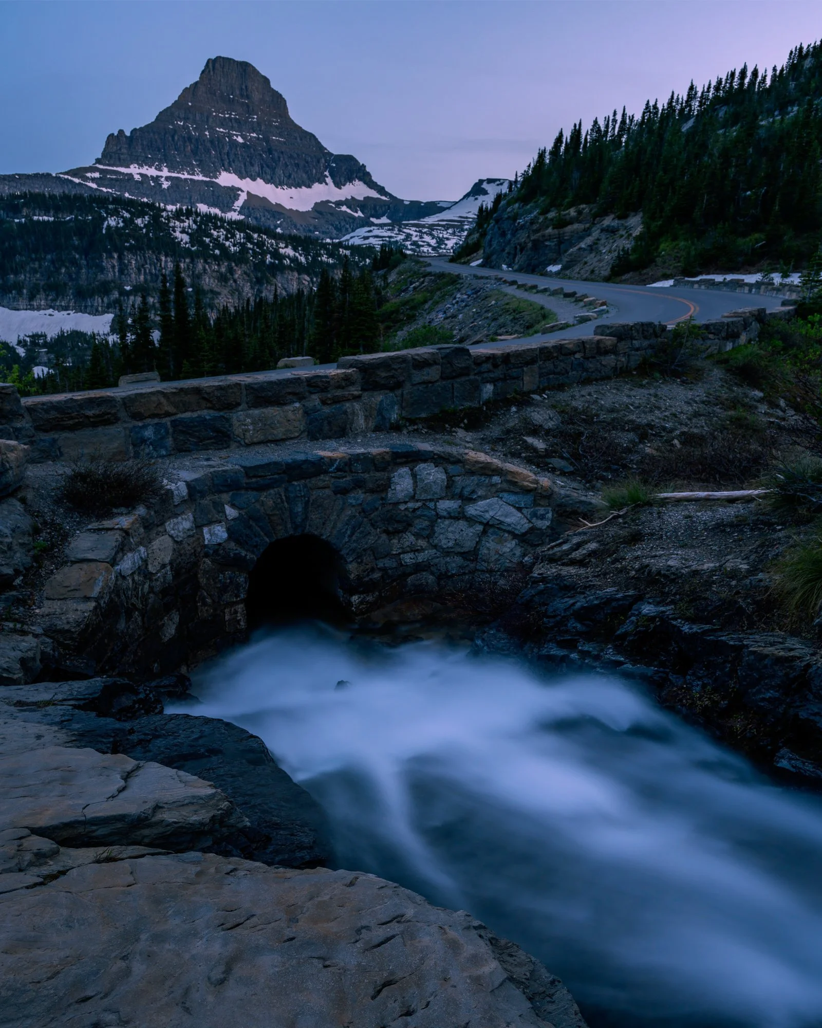 Mountain landscape with a winding road, stone bridge over rushing water, pine trees, and snow-capped peaks at dusk.