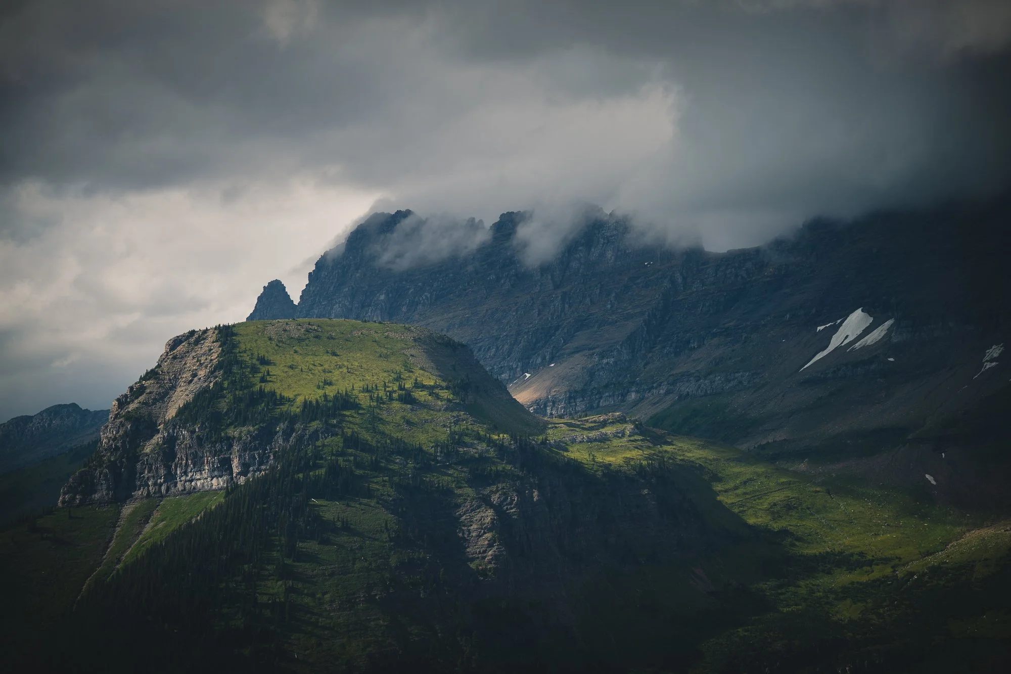 Cloudy mountain landscape with lush green hills and rugged peaks shrouded in mist and clouds.