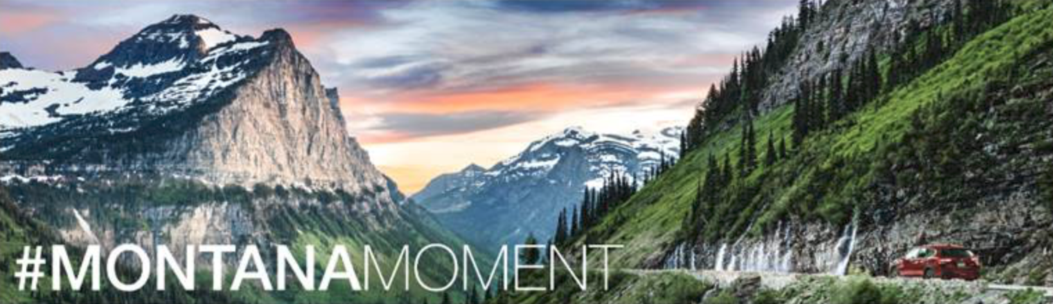 Scenic view of a mountain valley with snow-capped peaks, lush green hills, and a winding road with a red car, during sunset or sunrise, with the text '#MONTANA MOMENT' overlaid.