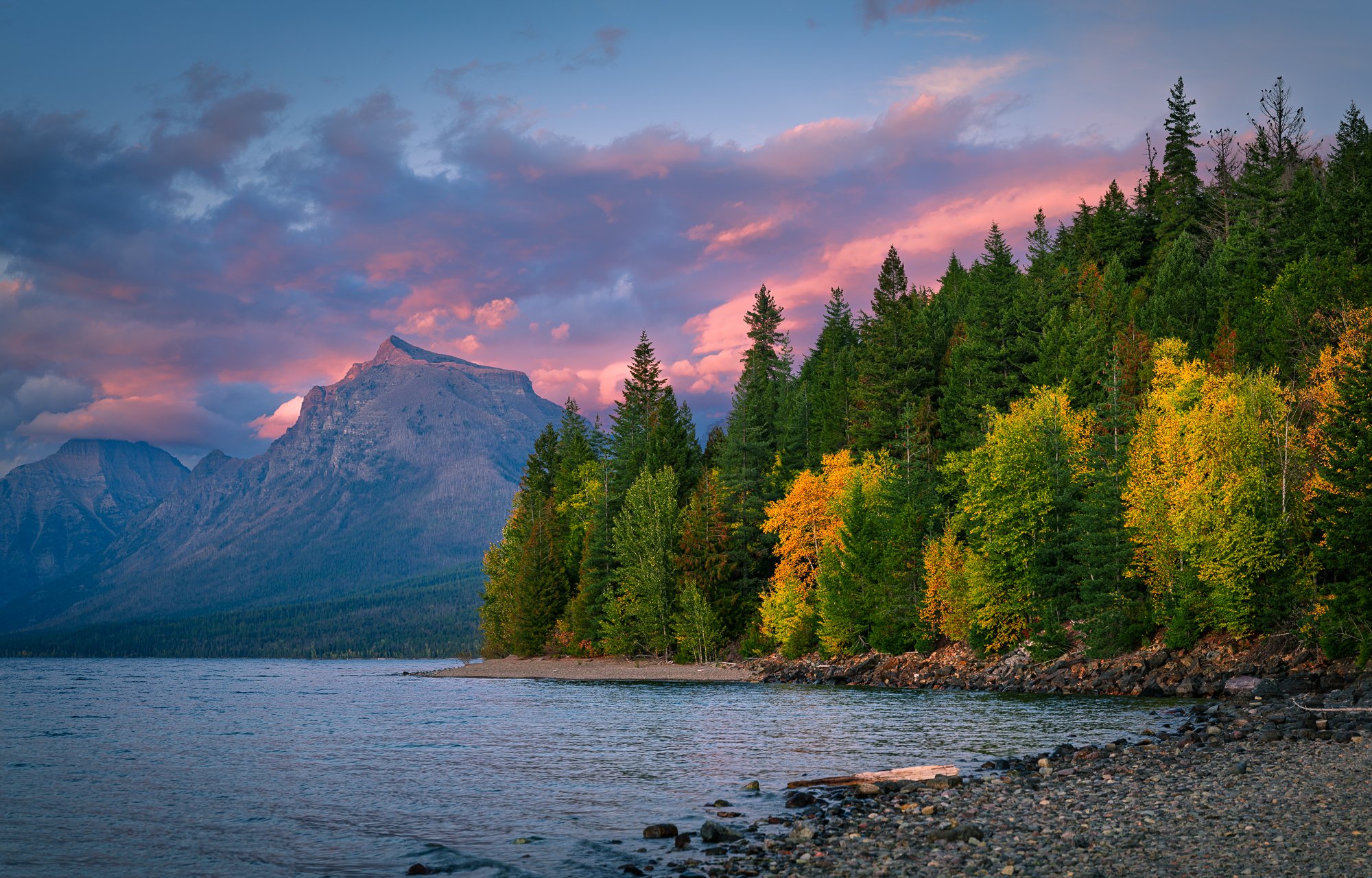 A scenic view of a lake with a rocky shoreline, surrounded by colorful trees in fall, with mountains in the background and a sky with pink and purple clouds at sunset.
