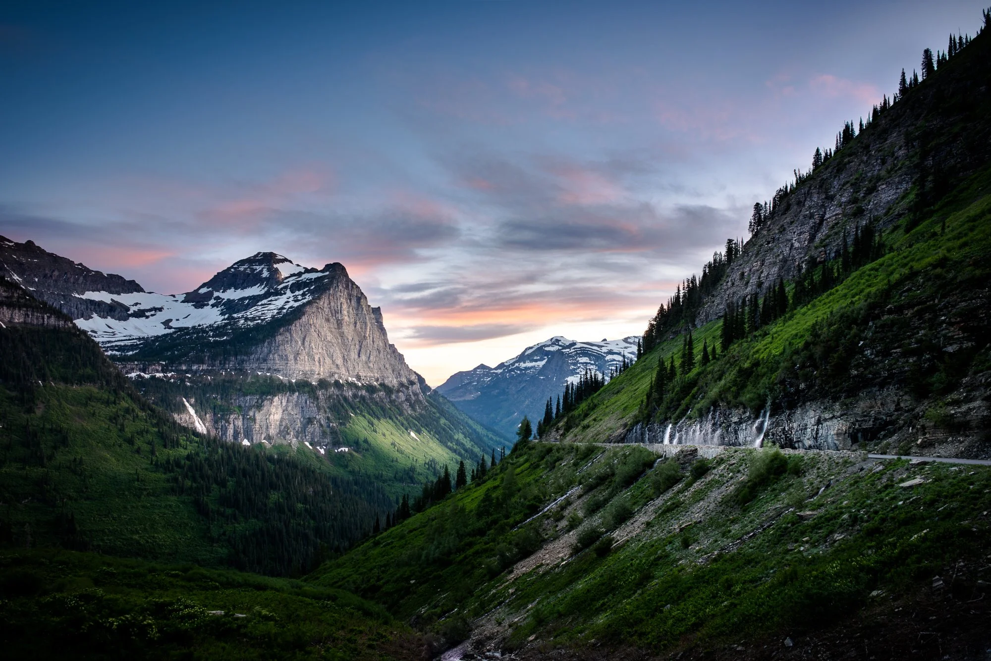 A scenic mountain valley with snow-capped peaks, green slopes, and a colorful sunset sky.