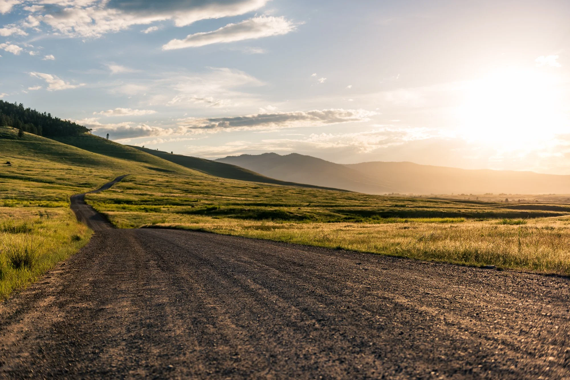 Scenic landscape of a winding gravel road through rolling green hills with mountains in the distance, under a partly cloudy sky at sunset.