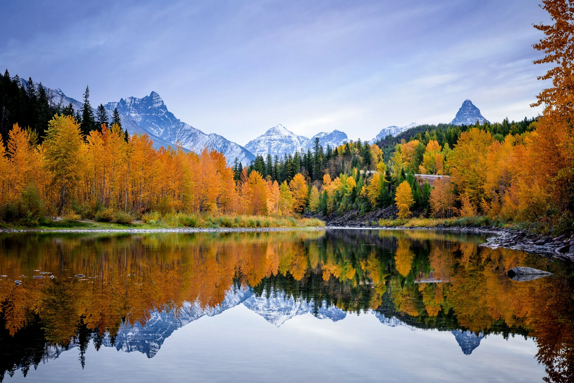 Mountains with snow caps behind a forest of autumn-colored trees and a calm river reflecting the landscape.