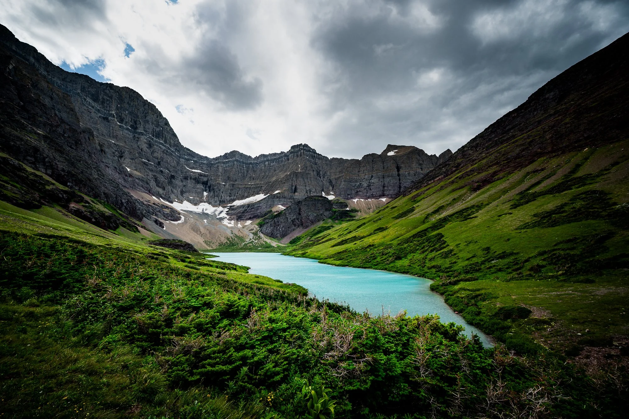Mountain valley with a turquoise lake, green slopes, and rugged cliffs under a cloudy sky.