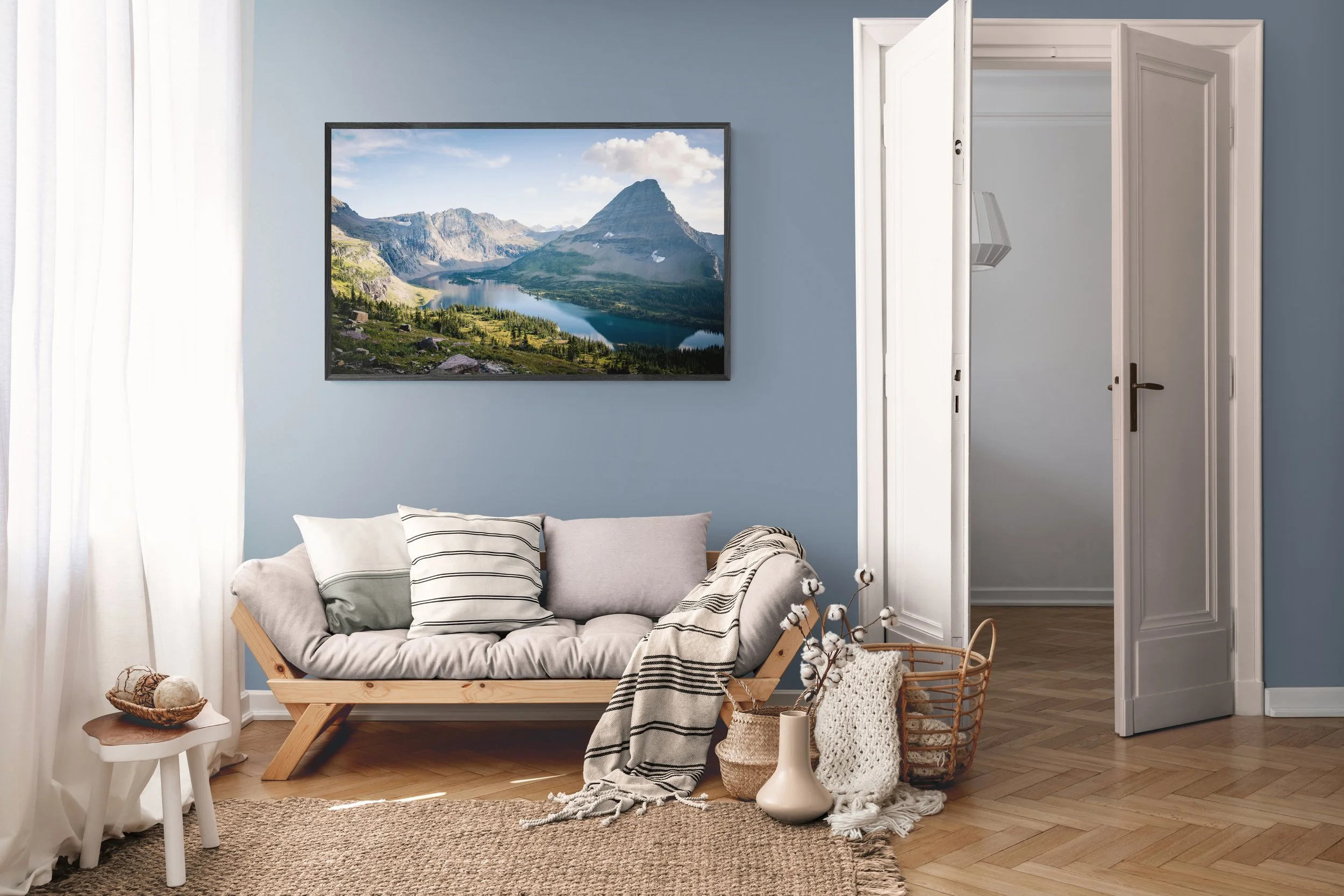 Living room with light blue walls, a framed landscape photograph, a beige sofa with striped and plain pillows, a small white side table with decorative items, a basket with cotton branches, and an open closet door.