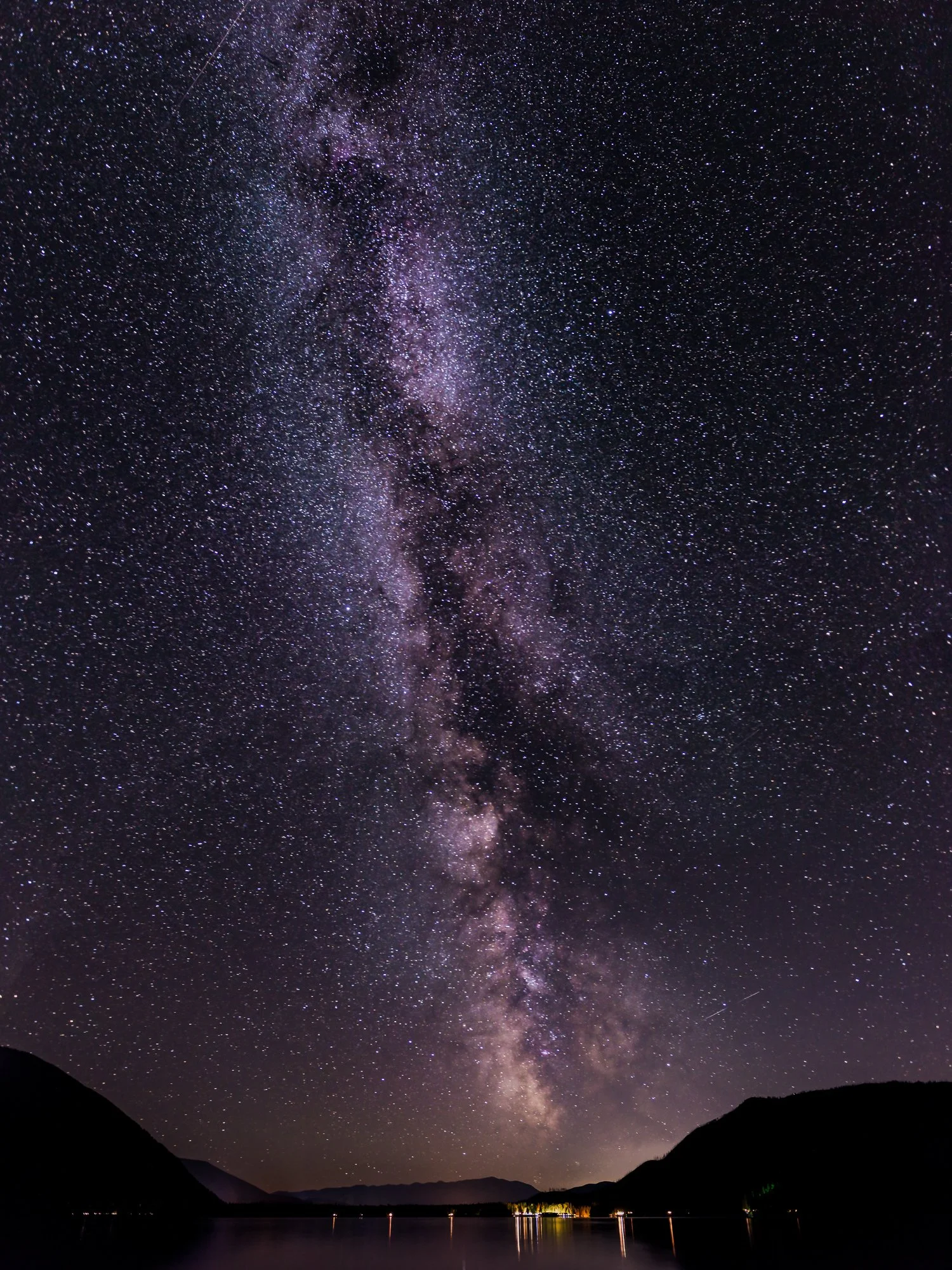 Night sky filled with countless stars and the Milky Way galaxy visible, with a dark silhouette of mountains and a calm lake below.