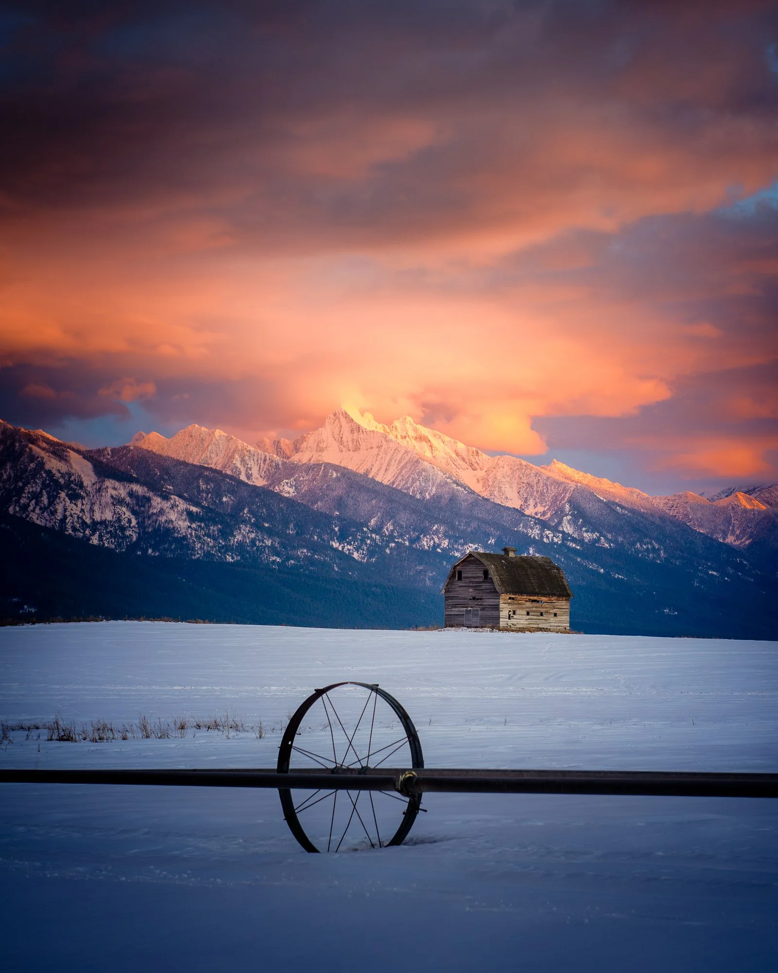 Snow-covered field with an old wooden barn and a mountain range in the background at sunset, with a snow-covered wheel in the foreground and colorful clouds in the sky.