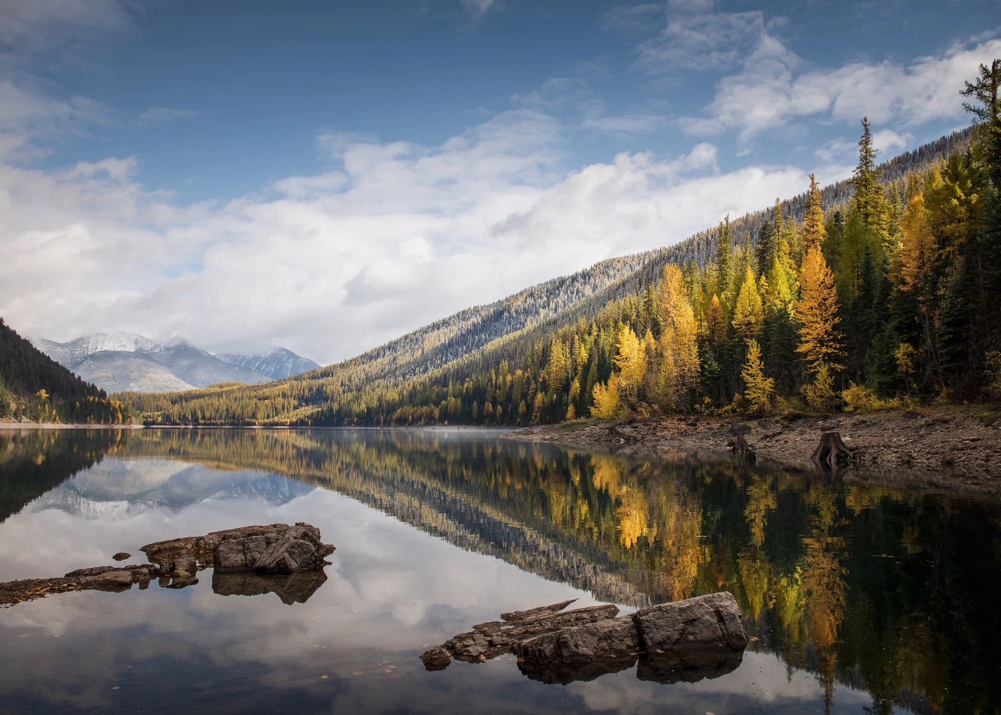A calm mountain lake reflecting the forested mountainside and blue sky with clouds.