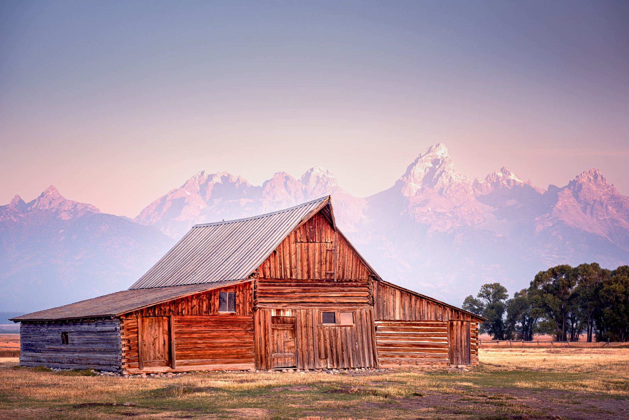 A rustic wooden barn with a metal roof in an open field with mountains in the background during sunset.