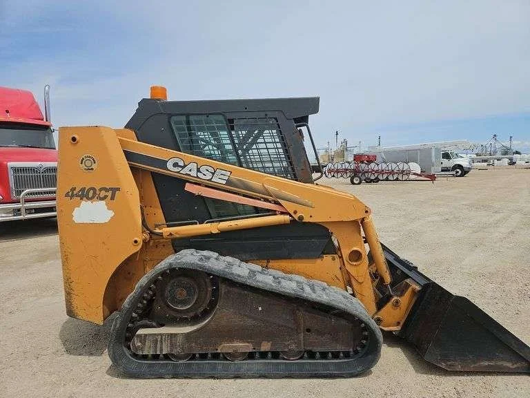 Yellow CASE 440CT compact track loader with a black bucket attachment, parked on a dirt lot with a red truck, white vehicles, and farm equipment in the background.