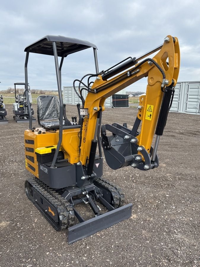 Small yellow and black mini excavator with rubber tracks and a cab, parked outdoors on a gravel surface with industrial containers and other equipment in the background.