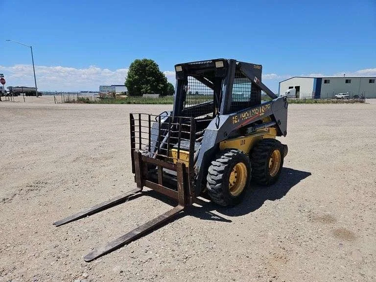 A small skid-steer loader with a fork attachment on a dirt lot.