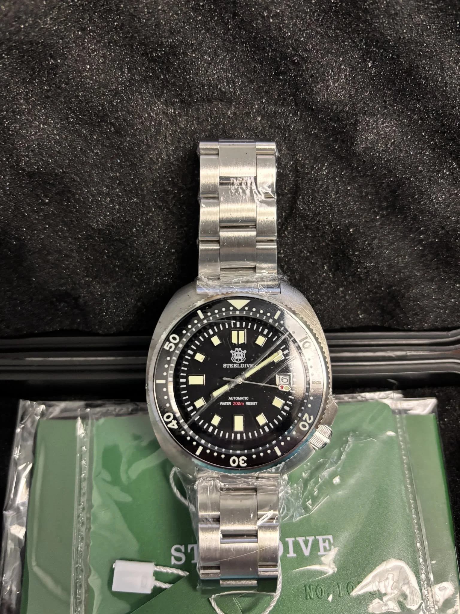A stainless steel wristwatch with a black dial and yellow markings, showing a date of the 16th, resting on a green surface with plastic wrapping.