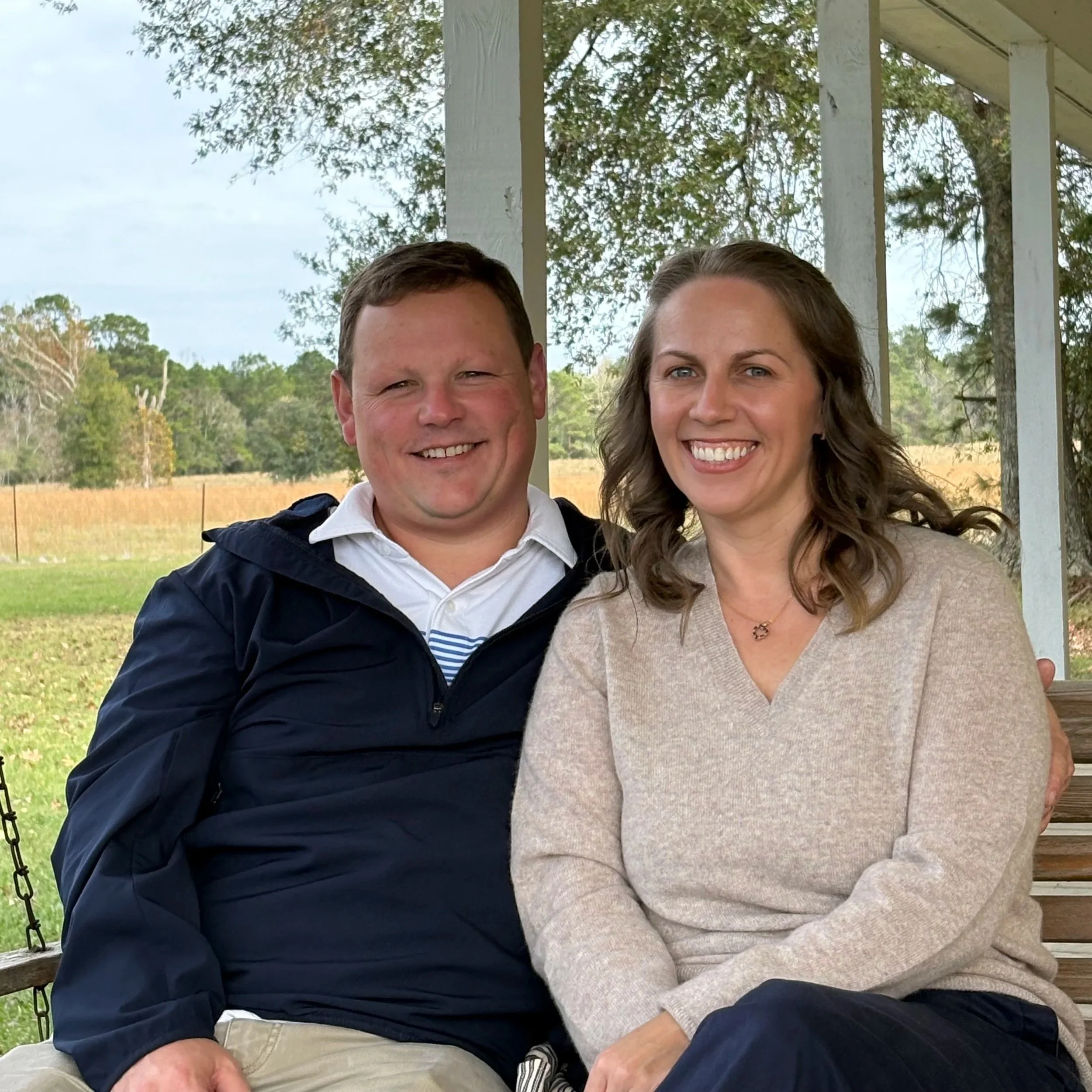 A smiling couple sitting on a wooden porch in a rural setting, with trees and open field in the background.