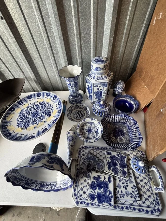 Collection of blue and white ceramic dishes, bowls, vases, and decorative items on a white table, with a cardboard box on the right and a corrugated metal wall in the background.