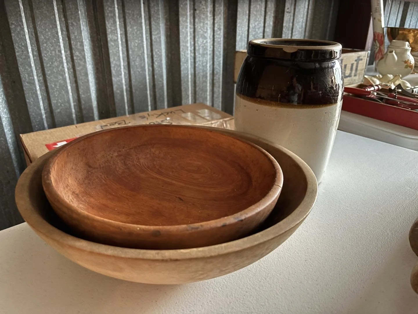 Set of nested wooden bowls and a ceramic jar on a white surface, with a metal wall background and decorative items in a tray.