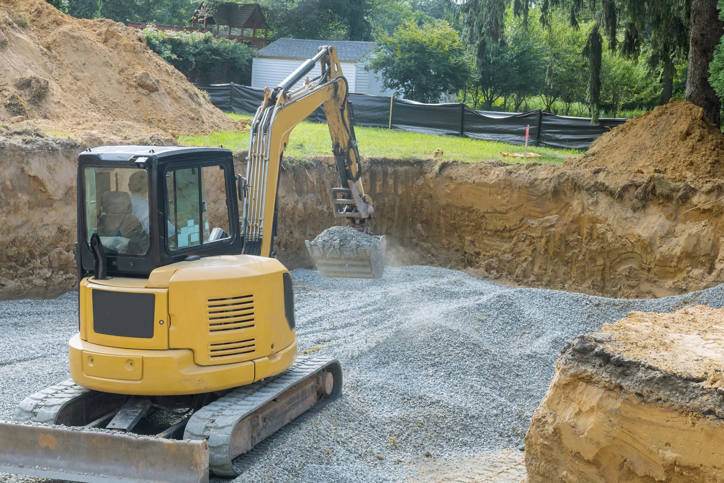 A yellow compact excavator moving gravel at a construction site in a grassy area with trees and houses in the background.