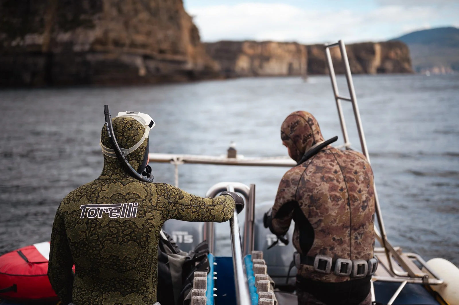 Two people in camouflage wetsuits on a boat, looking out at the water and cliffs in the distance, preparing for a water activity.