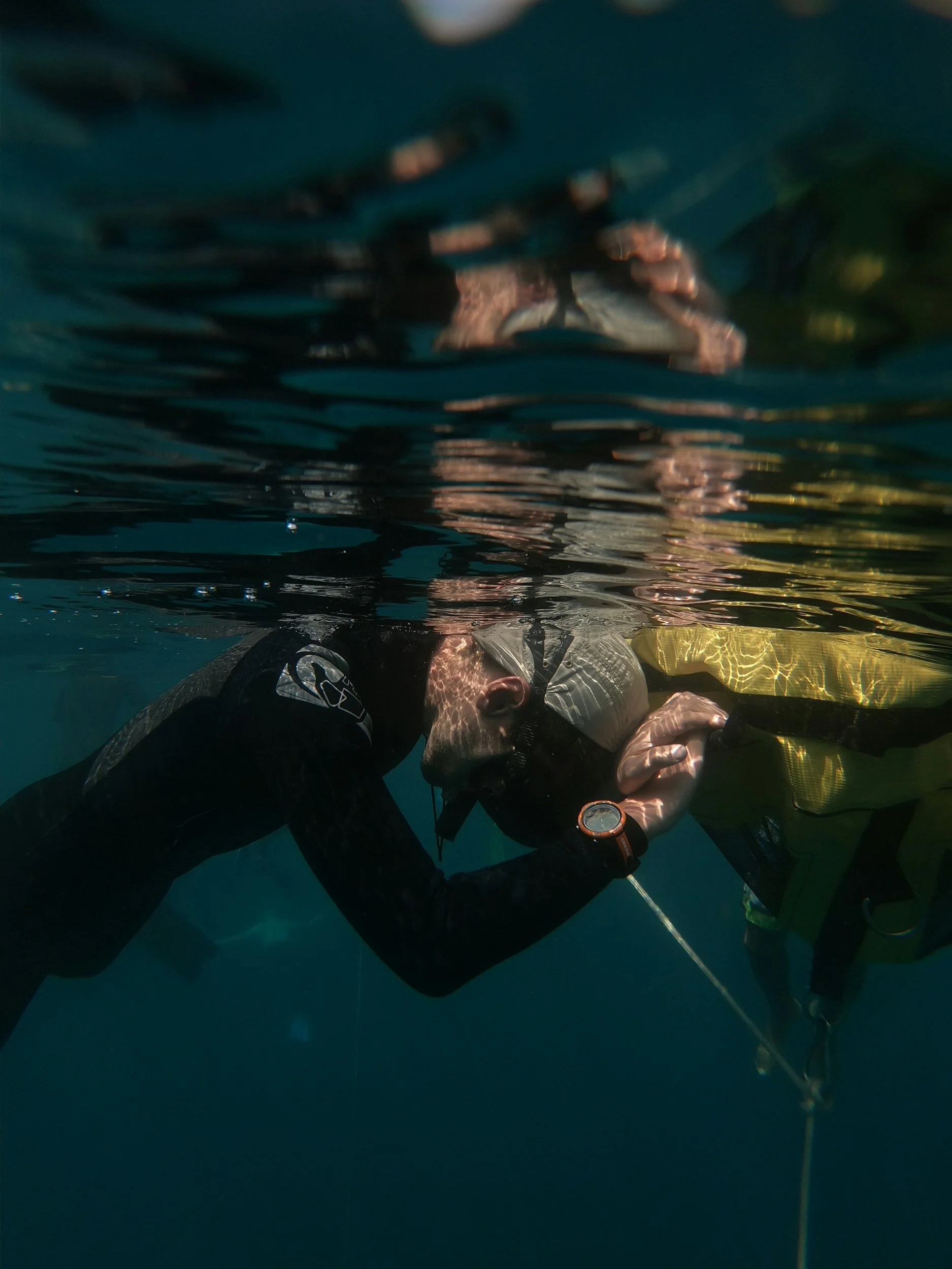 A person in scuba gear underwater, it appears they are conducting a rescue or assisting another diver, with one diver's face visible, wearing a mask, snorkel, wetsuit, and a yellow buoyancy vest, using a communication or safety device.